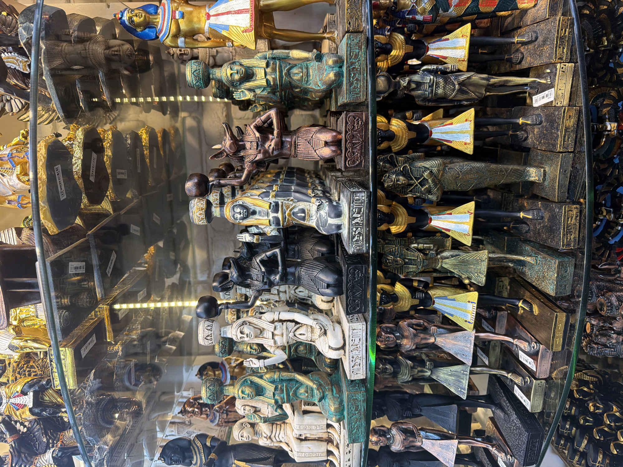This image depicts a shelf display of Egyptian souvenirs in a shop in Sharm El Sheikh, Egypt. The display is organized in rows on glass shelves, showcasing numerous statuettes and figures. The subjects are primarily Egyptian deities and pharaohs, rendered in various materials such as bronze, stone, and painted resin. Prominent figures include representations of Anubis, Bastet, and various pharaonic figures in traditional attire and headdresses. Many of the figures are detailed and appear to be decorative items sold in a tourist market. The shelves themselves are reflective, showing the items on shelves above and below. The lighting appears to be artificial, possibly from overhead fluorescent bulbs. The overall environment suggests a retail setting focused on Egyptian cultural artifacts.