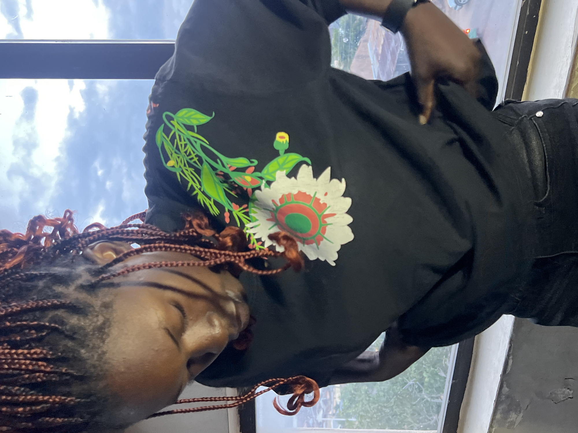 A dark-skinned woman with braided hair stands in front of a window in Jos, Nigeria, looking down and adjusting her black t-shirt which features a floral design. The t-shirt has a large white flower with red and green accents on the chest, and a green vine with leaves extending from the upper right side of the flower. She wears dark denim jeans and a black smartwatch. The background outside the window shows a cloudy sky and a glimpse of rooftops and trees, suggesting an urban environment. The interior setting appears to be a room with a concrete ledge beneath the window.