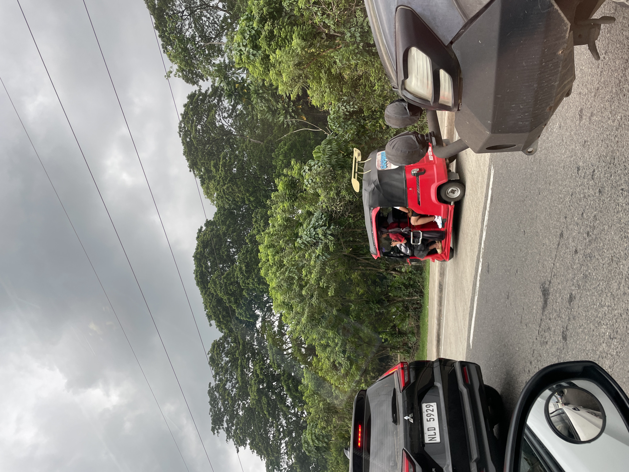 The image shows a red motorized tricycle with passengers traveling on a paved road in a rural area of Davao City, Philippines. A black Mitsubishi SUV with license plate NLD 5929 is behind the tricycle. The sky is overcast, and lush green trees and foliage are visible on the side of the road. Overhead power lines run alongside the road. The side mirror of a white vehicle is visible on the bottom right of the image, and a dark colored SUV or truck is positioned ahead.