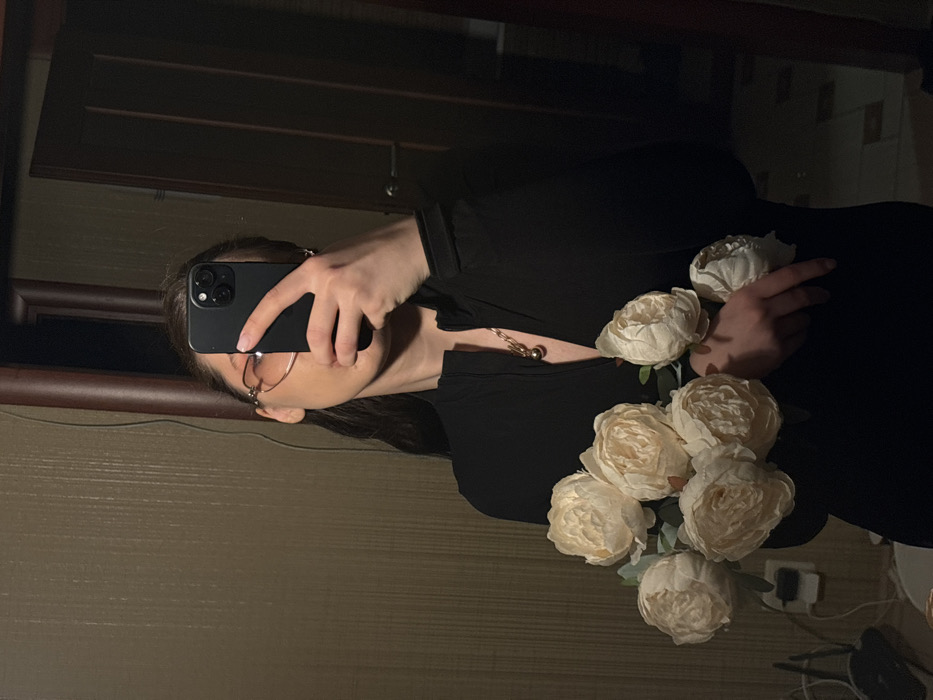 The image displays a mirror selfie of a person holding a bouquet of flowers. The photograph is oriented sideways, rotated 90 degrees clockwise.
In the center of the frame, a person is visible, primarily from the chest up. Their face is largely obscured by a dark-colored smartphone, which they are holding up to take the picture. The smartphone appears to be a modern model, possibly an iPhone, identifiable by its distinct camera bump with three lenses on the back. The person is wearing eyeglasses with thin, possibly gold or rose-gold colored frames, and their dark hair is visible around their face.
They are dressed in a dark, likely black, long-sleeved top or dress. A prominent gold-toned necklace with a spherical or circular pendant hangs around their neck and rests on their chest. Their right hand is cradling a bouquet of artificial flowers, which resemble peonies or large roses. The flowers are a uniform light, creamy white or very pale yellow color, and there are approximately six to seven visible blooms with green stems and leaves. The person's nails on both hands appear unpainted.
The background, which is the reflection in the mirror, is dimly lit. To the top left (if the image were upright), there is a dark, possibly wooden or wood-paneled surface, which could be part of a furniture piece or wall decor. The wall behind the person's lower body (bottom right if upright) has a lighter, textured appearance, possibly wallpaper or a painted surface with subtle horizontal lines. In the very bottom right corner of the original image (which would be the lower part of the reflected room if upright), a white electrical outlet or device is partially visible on the floor or a low surface, along with some dark wires or cables.
The overall lighting of the scene is low, creating a somewhat intimate and subdued atmosphere with soft shadows. No text is visible in the image.