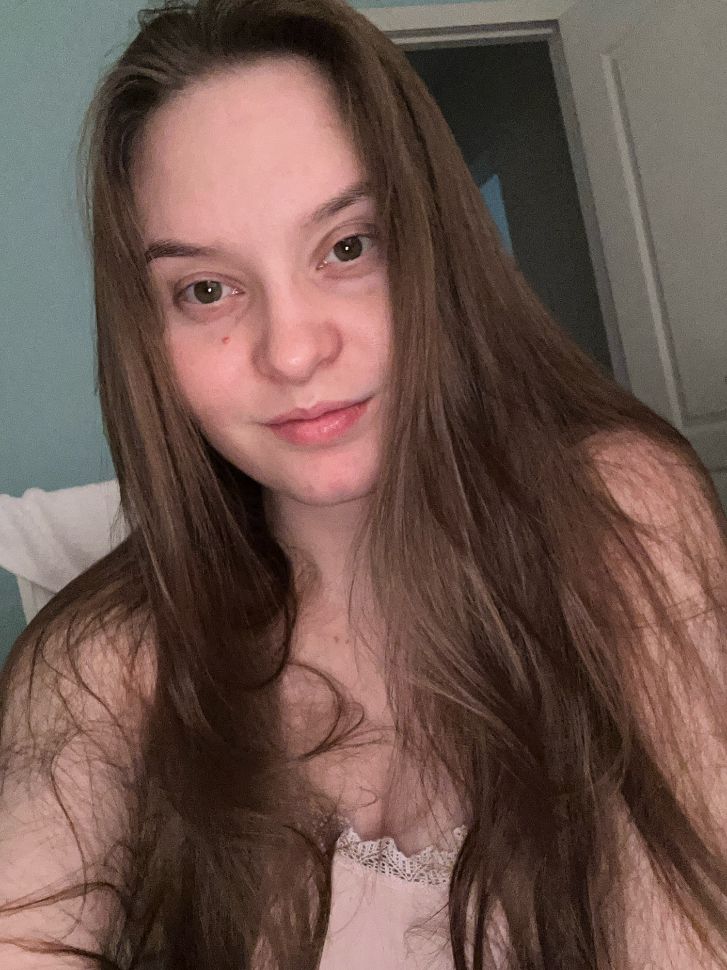 A young woman with long, dark brown hair is shown in a close-up selfie. She is looking directly at the camera with a neutral expression. Her hair falls loosely around her shoulders and chest. She is wearing a light-colored garment with lace detailing visible at the neckline. The background appears to be an indoor setting, with a pale blue wall on the left and a doorway with a white door on the right. The lighting is soft and ambient. There are no discernible objects or interactions to note, and the image does not contain information about the city of Noviy, Russia.