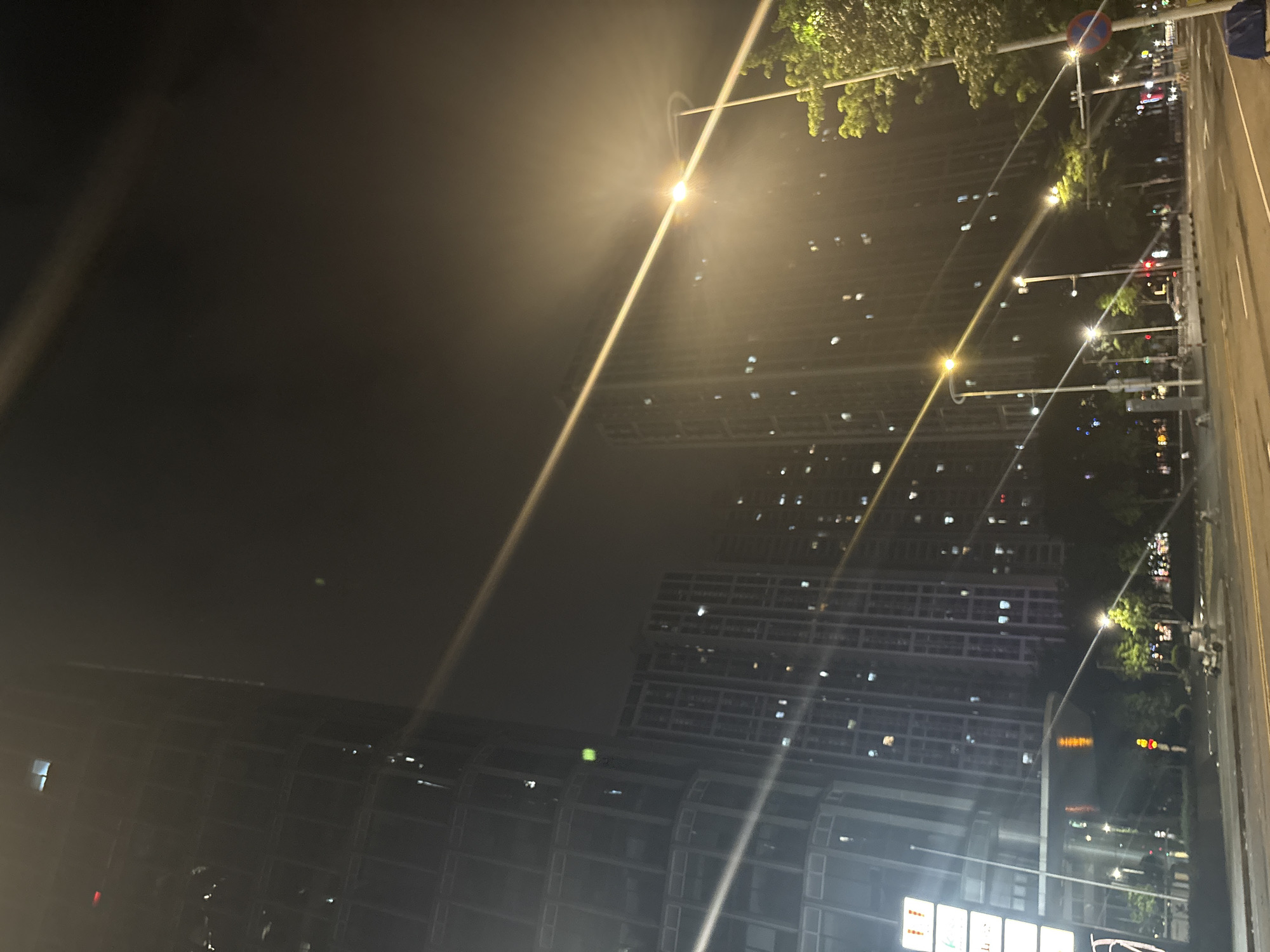 This media file captures a nighttime scene in Haizhu District, China, likely from a high vantage point. The foreground is dominated by the dark, blurred shapes of modern buildings, illuminated by scattered lights from their windows. Some of these buildings appear to be under construction or are in a state of significant development, with visible scaffolding and structural elements.
Streaks of light from street lamps and possibly vehicle headlights cut diagonally across the frame, suggesting motion or a long exposure. In the midground and background, more illuminated high-rise buildings are visible, creating a dense urban landscape. There are also trees with green foliage visible, adding a touch of nature to the otherwise artificial environment. The sky is dark, indicating it is nighttime, and there appears to be some atmospheric haze or fog, which softens the lights and creates a slightly ethereal quality.
The overall mood is one of urban vibrancy and activity at night. There are no clearly identifiable people or specific activities taking place, but the presence of numerous lights suggests a populated area. A red traffic signal and a circular sign with red and blue markings are visible on a pole along a street, providing a clue to the location as a public thoroughfare. Some faint text might be present on distant signs, but it is illegible due to the distance and low light conditions.