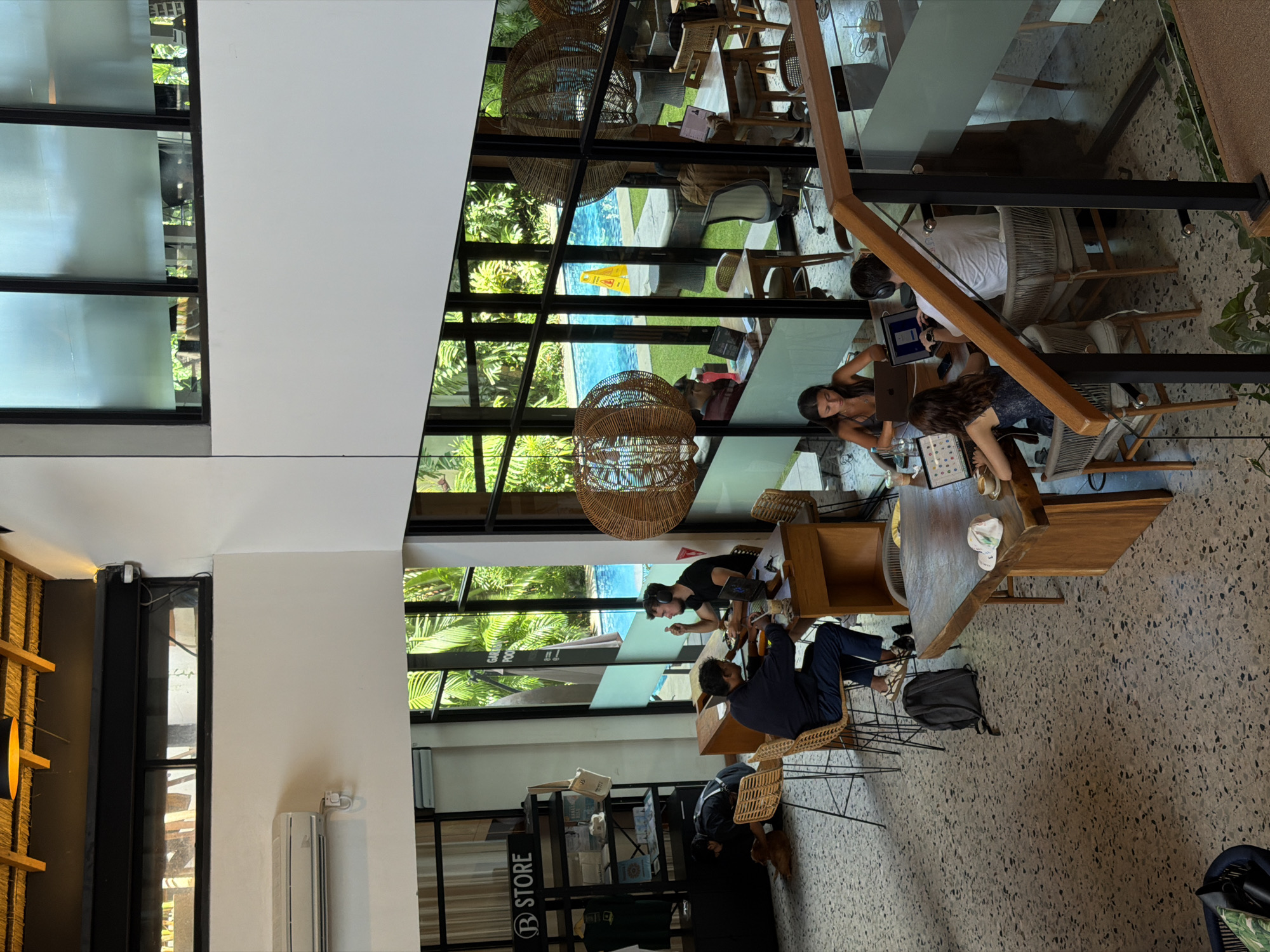 The image depicts an interior scene of a co-working space or café in North Kuta, Indonesia. Sunlight streams through large windows, reflecting off a swimming pool outside. Several individuals are working at tables with laptops. One group is seated closely together near a staircase, while others are spread across the space. The interior design includes rattan light fixtures and a small retail display labeled "Store." The flooring is terrazzo.
