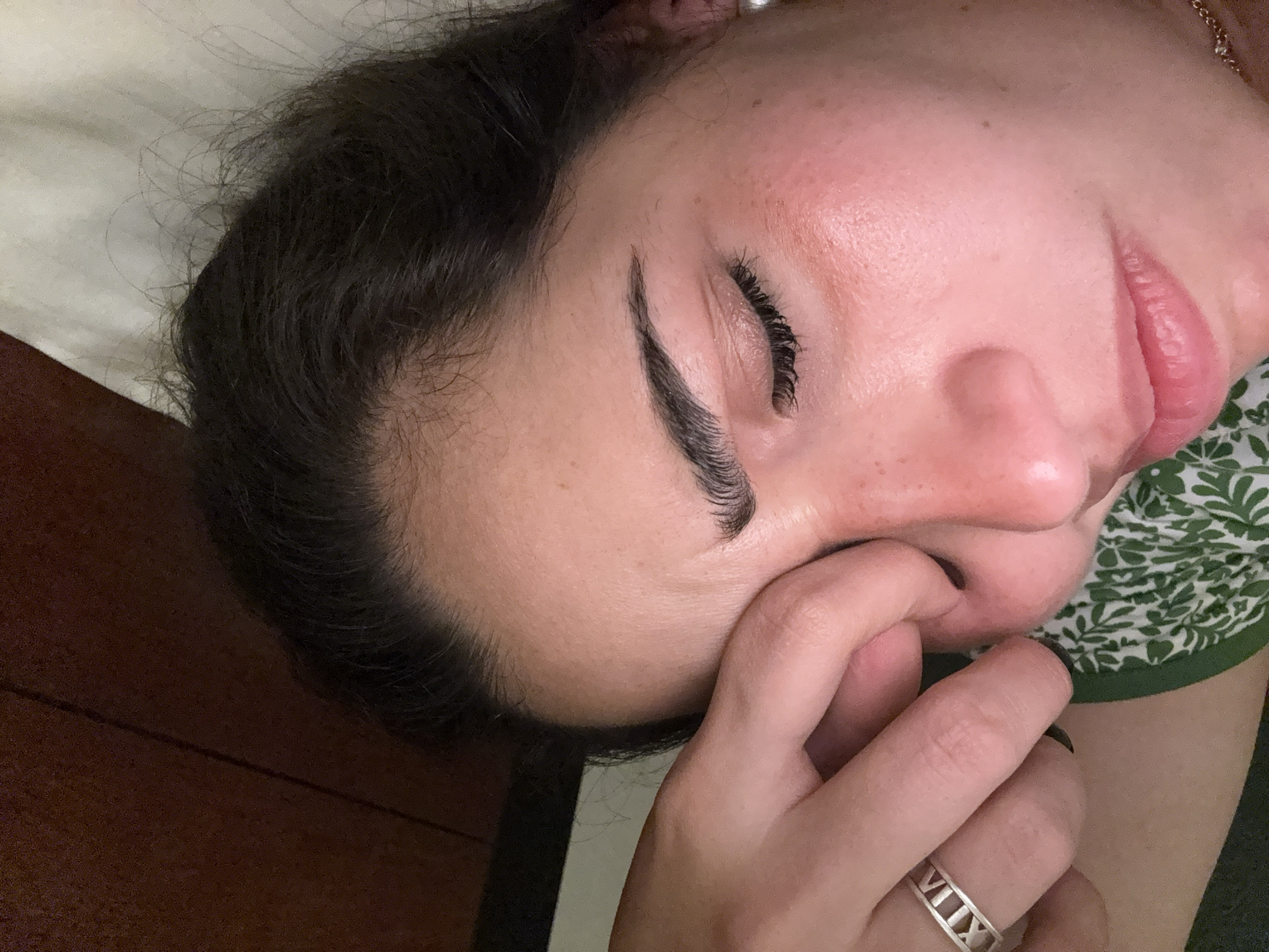 A close-up image shows a person lying down, possibly on a bed or treatment table. The person appears to be female with dark hair, closed eyes with lash extensions, and well-defined eyebrows. She is wearing a ring on her finger with Roman numerals. Her hand is near her face, touching the nose. The background includes a patterned fabric, possibly part of a garment. The location is indeterminable.