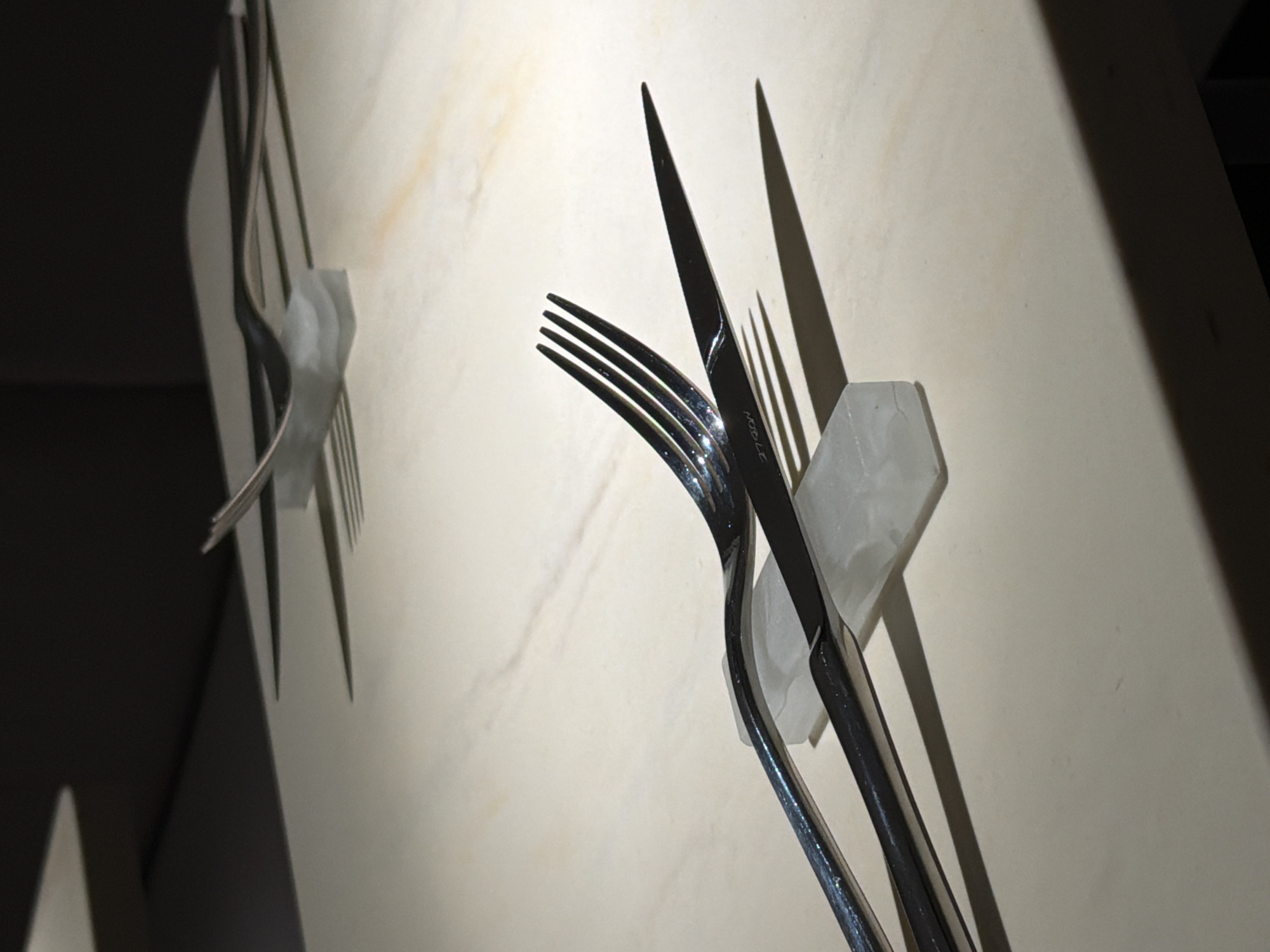 A low-angle shot depicts two sets of stainless steel forks and knives displayed on a pale, marble-textured wall, appearing to be suspended by white, hexagonal holders. The first set, in the background, consists of a fork slightly out of focus. The second, foreground set consists of a fork and knife. The knife blade is visible and the knife's brand, "Noble," is visible. Shadows cast by the cutlery accentuate their shapes on the wall. The scene appears to be indoors, possibly in a dining or culinary setting. There's insufficient information to confirm a location in Moscow, Russia.