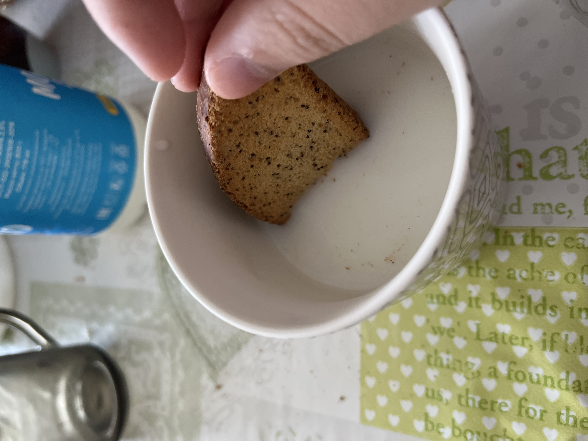 A person's hand is holding a square-shaped piece of biscotti, partially submerged in milk within a white bowl. The biscotti is golden brown with visible poppy seeds or similar small dark particles. The bowl has a decorative pattern around its rim. In the background, a blue carton with text and a glass mug are visible, along with a patterned tablecloth. The scene appears to be indoors, likely a kitchen or dining area, on a table. The lighting is natural, coming from the top right.