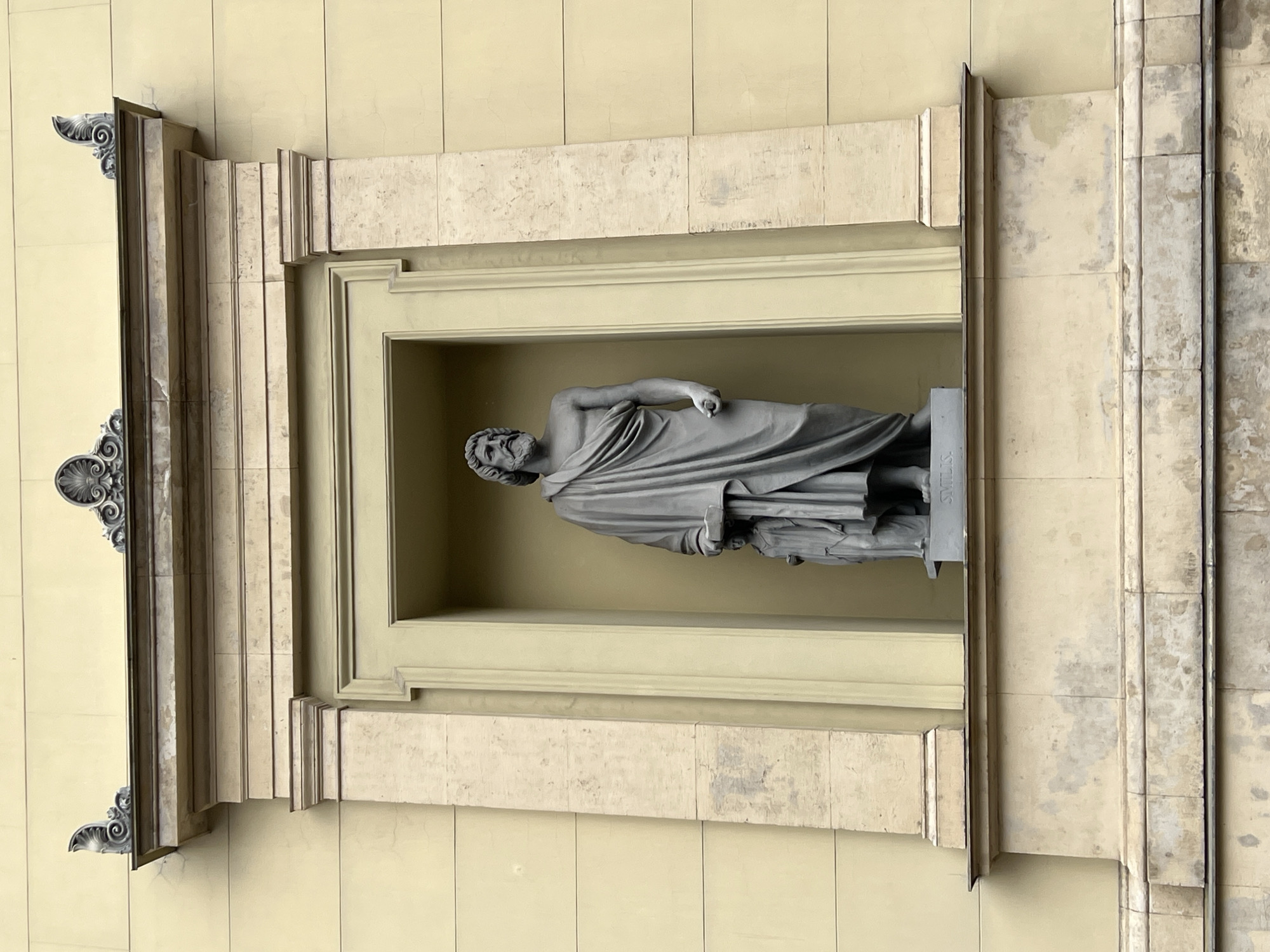 The image shows a niche in the facade of a building, likely in Saint Petersburg, Russia. Within the niche stands a grey stone statue of a bearded man in a toga, holding a scroll or book. To the left of the main statue, a smaller female figure stands. The niche is framed by an architectural element with decorative carvings at the top, and the building itself is a pale yellow color. Below the niche, the building's facade is made of stone blocks, showing signs of weathering and age. The statue is mounted on a grey plinth, with some lettering visible. The overall impression is of a classical architectural detail on an old building.
