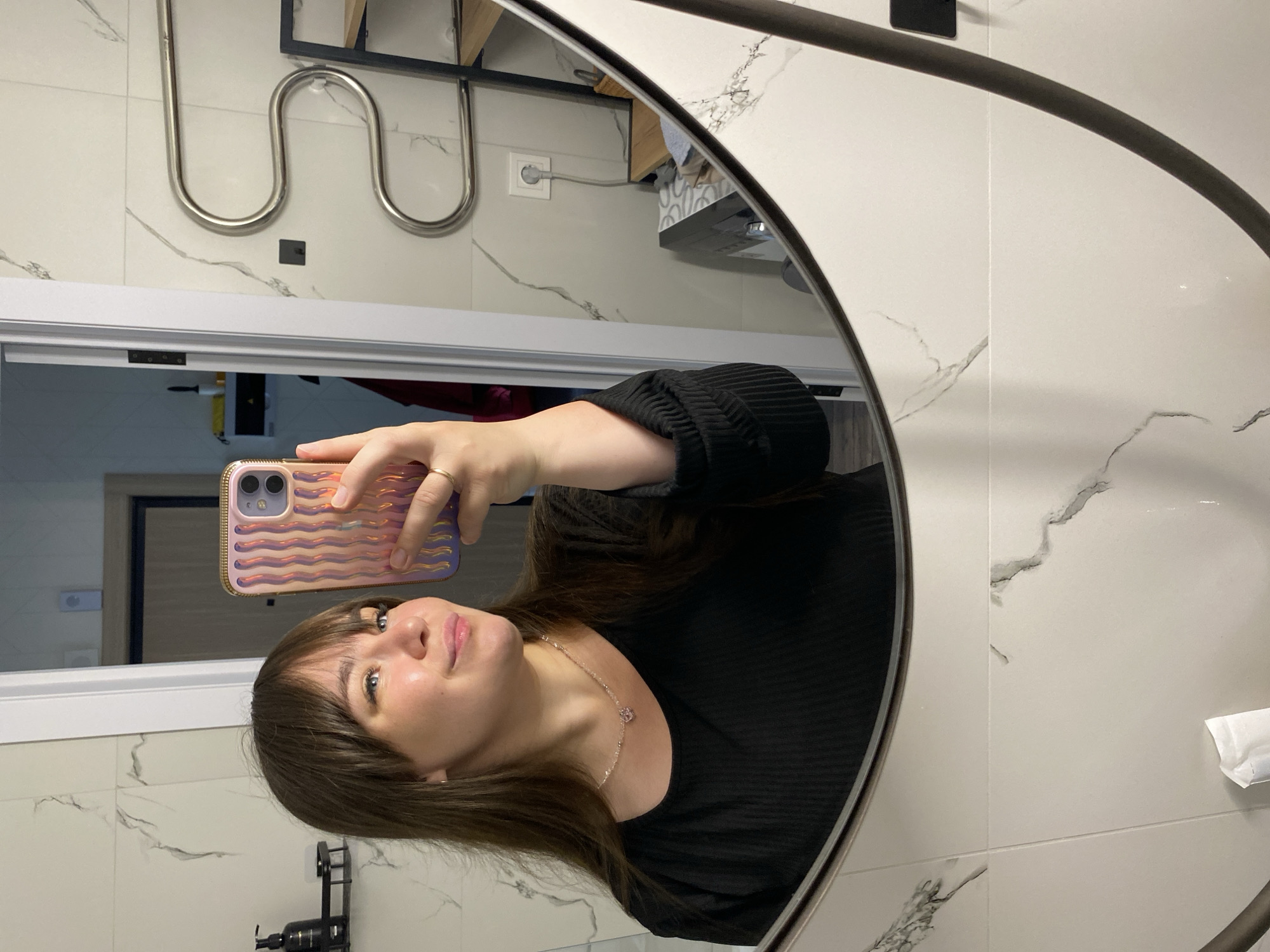A woman is taking a mirror selfie in what appears to be a modern bathroom. She is positioned to the left side of a large, dark-framed mirror, holding a light pink/purple smartphone with a wavy, iridescent case up to capture her reflection. The phone has two visible camera lenses. The woman has long brown hair with bangs, is wearing a black, ribbed long-sleeved top, and a delicate necklace with a small pendant. She is looking upwards towards the phone, with a neutral to slightly pleasant expression.
The reflected background behind her reveals a white tiled wall with gray marble-like veining. Visible elements in the reflection include a chrome-colored, S-shaped towel warmer or radiator mounted on the wall, a white power outlet, and a partially visible wooden shelf or cabinet to the right. A dark, possibly black, dispenser bottle is seen in the bottom left of the reflection. The white door frame and a sliver of a room beyond it are visible in the upper left of the mirror. A small white crumpled tissue or paper is also seen on the wall to the right. The overall setting suggests an interior space, likely a bathroom, with contemporary finishes. There is no discernible text visible in the image.