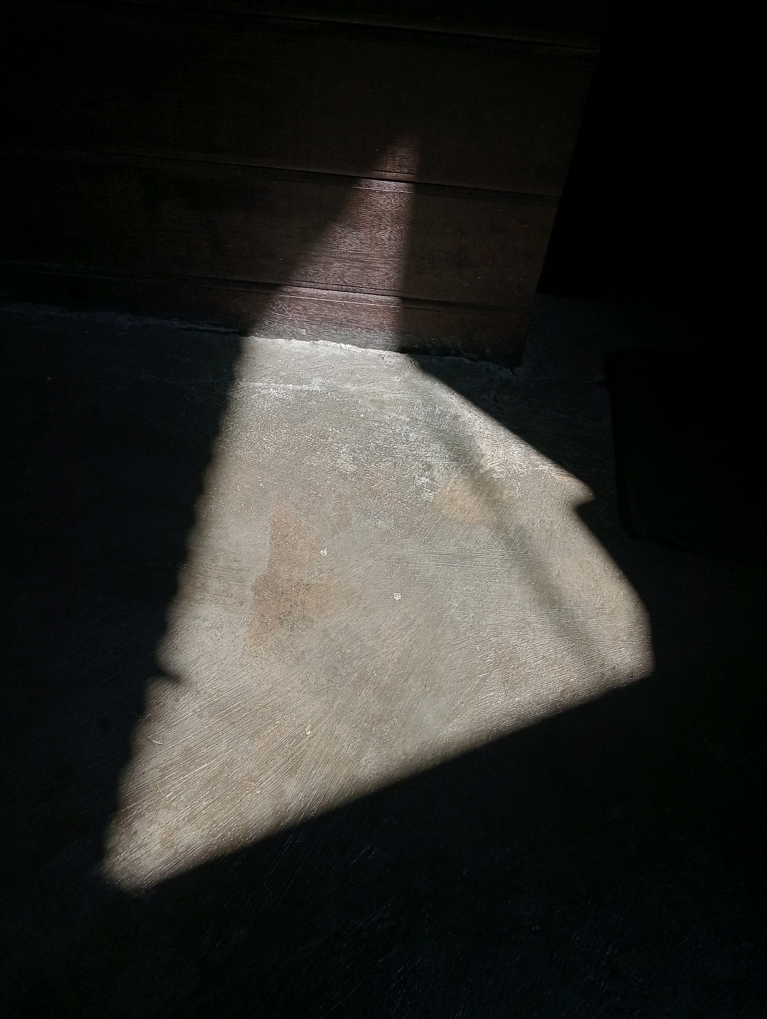 This image depicts a stark play of light and shadow on a textured concrete floor adjacent to a dark wooden surface. A sharply defined triangular shadow, cast by an unseen object or architectural element, dominates the lower half of the frame, tapering from a broad base on the left towards the right. The concrete surface exhibits subtle variations in color and texture, with some areas appearing lighter and more granular, while others are darker and smoother. The wooden surface, likely part of a wall or piece of furniture, is dark and horizontally striated, with a band of light illuminating a portion of its lower edge. The scene is devoid of any human presence or discernible activity, creating a sense of stillness and abstraction. The overall impression is one of minimalist composition, focusing on the interplay of light, shadow, and surface textures. The image was captured in the city of Samarinda, Indonesia, though no specific landmarks or indicators of its exact location within the city are visible.