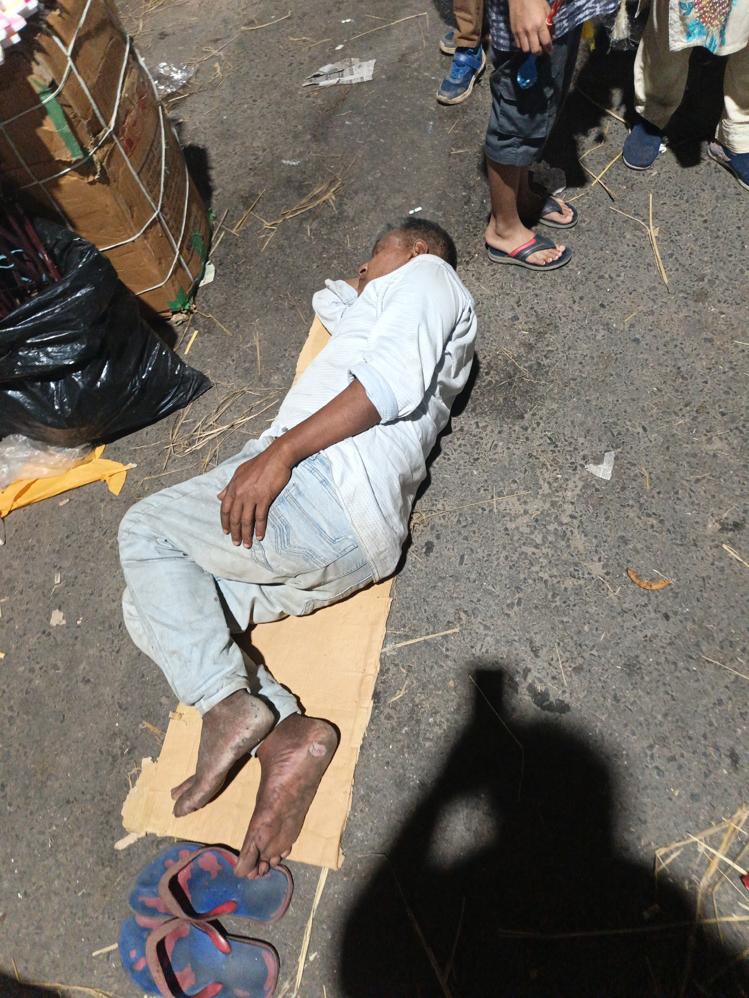 An adult male is lying on his side atop a piece of cardboard on a paved surface, likely a street. He is wearing a light-colored, button-down shirt and light-colored jeans. His feet are bare and appear dirty. A pair of blue and red flip-flops are next to his feet. To his left is a large, bound package and a black plastic bag with a piece of yellow fabric. The pavement is littered with straw and debris. Partial figures of several people standing nearby are visible, wearing casual clothing and sandals. The overall scene conveys the impression of poverty and possibly homelessness. The location is most likely Dhaka, Bangladesh.