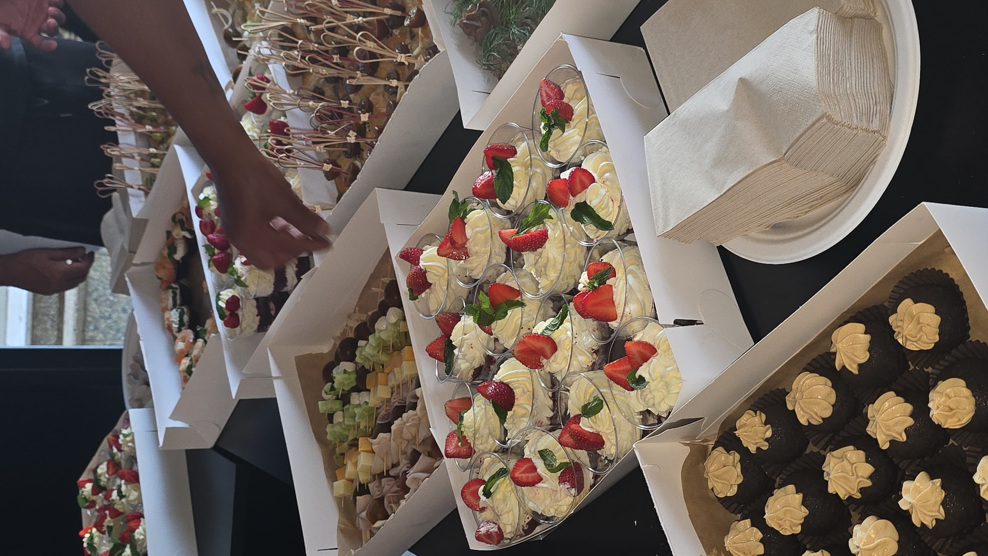 A table is laden with an assortment of catered desserts and appetizers, likely at an event in Trikomo, Northern Cyprus. In the foreground, a white cardboard box holds numerous small, dark chocolate desserts topped with a swirl of caramel-colored frosting. To the right of this box, a stack of light tan paper napkins rests on a white paper plate. Behind the chocolate desserts, several clear plastic cups contain layered desserts with whipped cream and sliced strawberries, garnished with mint leaves. Further back, a white cardboard box is filled with skewers of cheese cubes, grapes, and green melon balls. Beyond that, another white box holds appetizers on skewers, possibly involving olives and other small food items. In the background, a person's arm and hand are visible, reaching towards the desserts. The overall setting appears to be indoors, with a dark surface underneath the food displays.