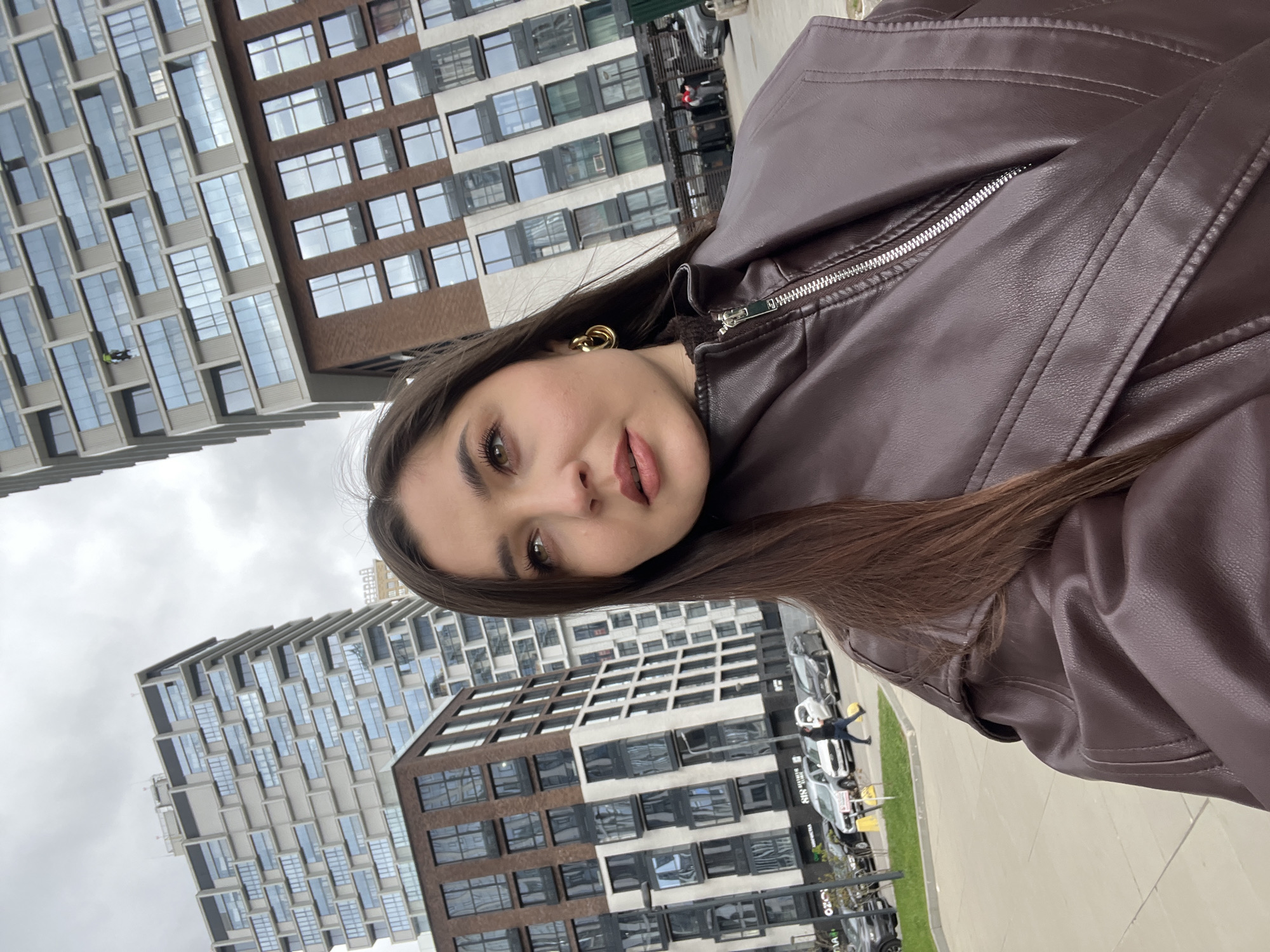 A young woman with long brown hair stands in front of modern buildings. She is wearing a brown leather jacket and gold earrings. The buildings are characterized by a grid-like pattern of windows. In the background, there are cars parked on the street and a person walking. This image appears to be taken in Khimki, Russia.