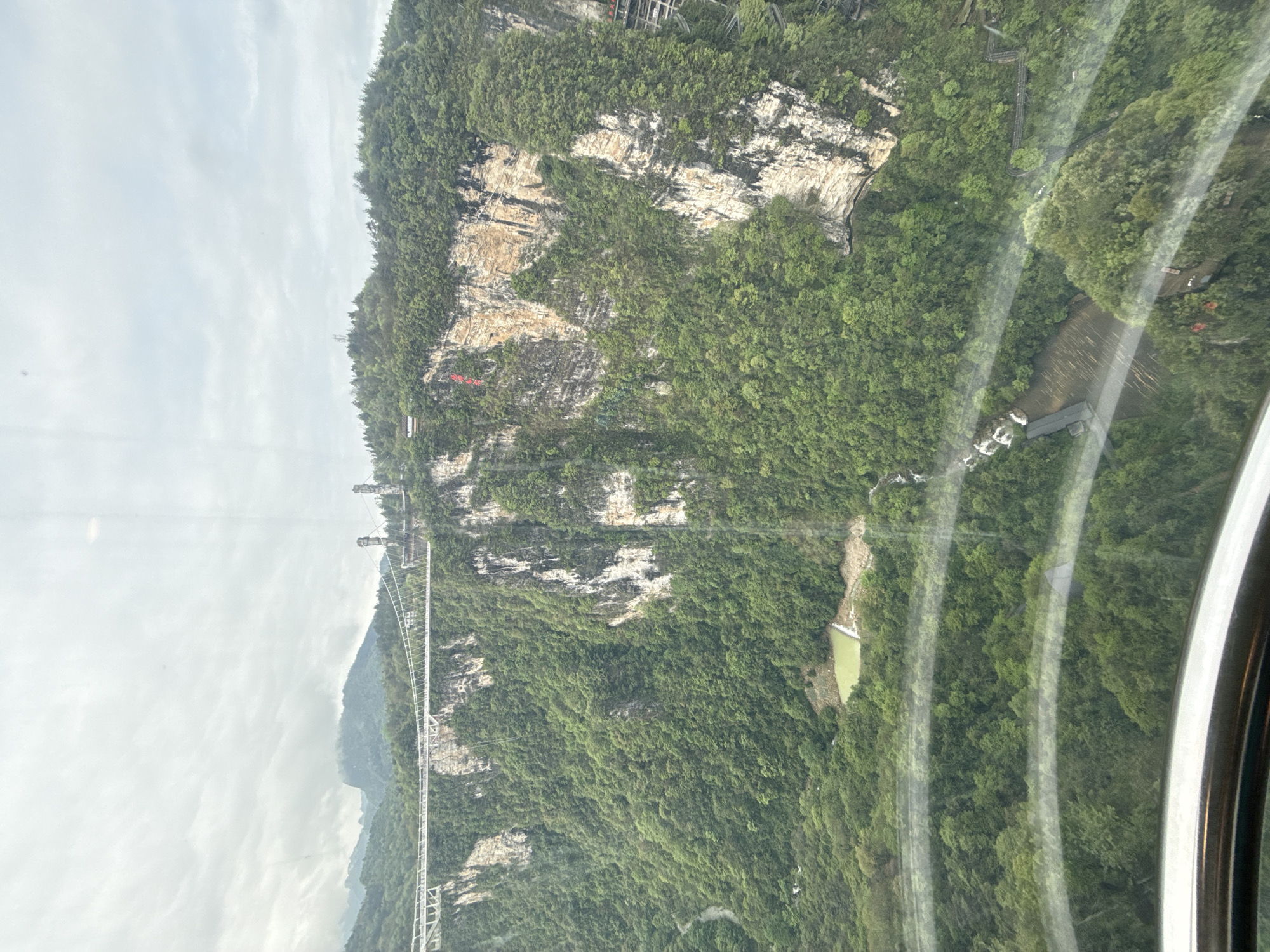 The image shows a landscape, possibly taken from inside a vehicle with the window visible on the right. The scene features a towering, green, tree-covered mountain with notable rock formations. A long, white suspension bridge spans across a portion of the landscape. A small pond or body of water is visible nestled among the trees. The setting appears to be the Zhangjiajie Glass Bridge in the city of Zhangjiajie, China.
