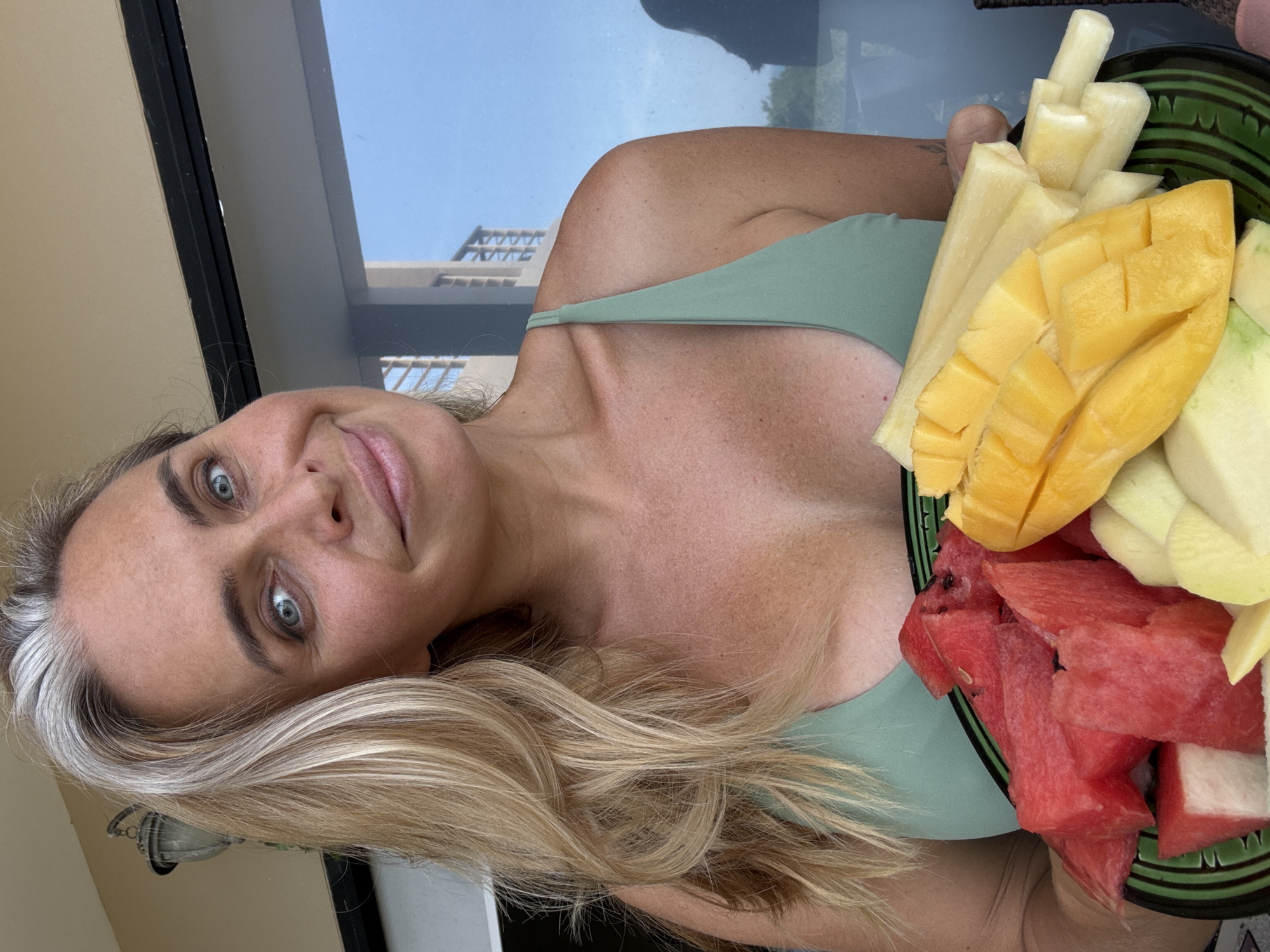 A fair-skinned woman with blonde hair, featuring prominent streaks of grey, holds a dark green plate of cut fruit. The fruit consists of chopped watermelon, sliced mango, and what appears to be peeled pineapple. She is wearing a light green tank top or bikini top. The background suggests an outdoor setting, possibly a balcony or patio, with a large window or glass door reflecting the sky and buildings. The lighting indicates it is daytime. The woman's facial expression is a slight smile, and she is looking towards the camera.