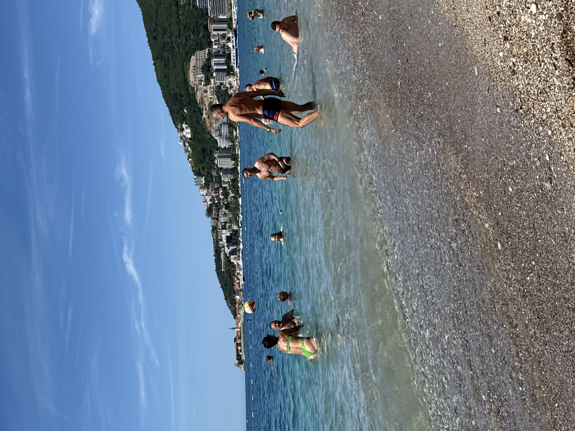 The image depicts a sunny day at a pebbly beach in Boreti, Montenegro. The foreground shows the shallow, clear, blue-green waters of the Adriatic Sea meeting a light brown and grey pebbly beach.
In the midground, numerous individuals are in the water. On the left, a woman in a bright green bikini is playing with a yellow, red, and white beach ball, splashing water. She is facing away from the camera, interacting with at least two other men in the water nearby. One man behind her is smiling, and another bald man is partially submerged to her right. Further to the right, a man with short grey hair and a beard, wearing striped swim shorts, stands facing right. Another man with a muscular build, in dark swim shorts, stands further left. To the far right, a man lies partially in the shallow water. Several other individuals are visible further out, either standing or swimming.
The background features a vast expanse of blue sea with white marker buoys. On the left, a historic fortified town with a prominent spire is visible on a peninsula, extending into the water. The coastline stretches to the right, dominated by a large, green, forested mountain covered with numerous white and light-colored buildings, indicating extensive urban development. Boats are anchored in the bay between the coastline and the distant town. The sky is a clear, bright blue with a few faint, wispy clouds.