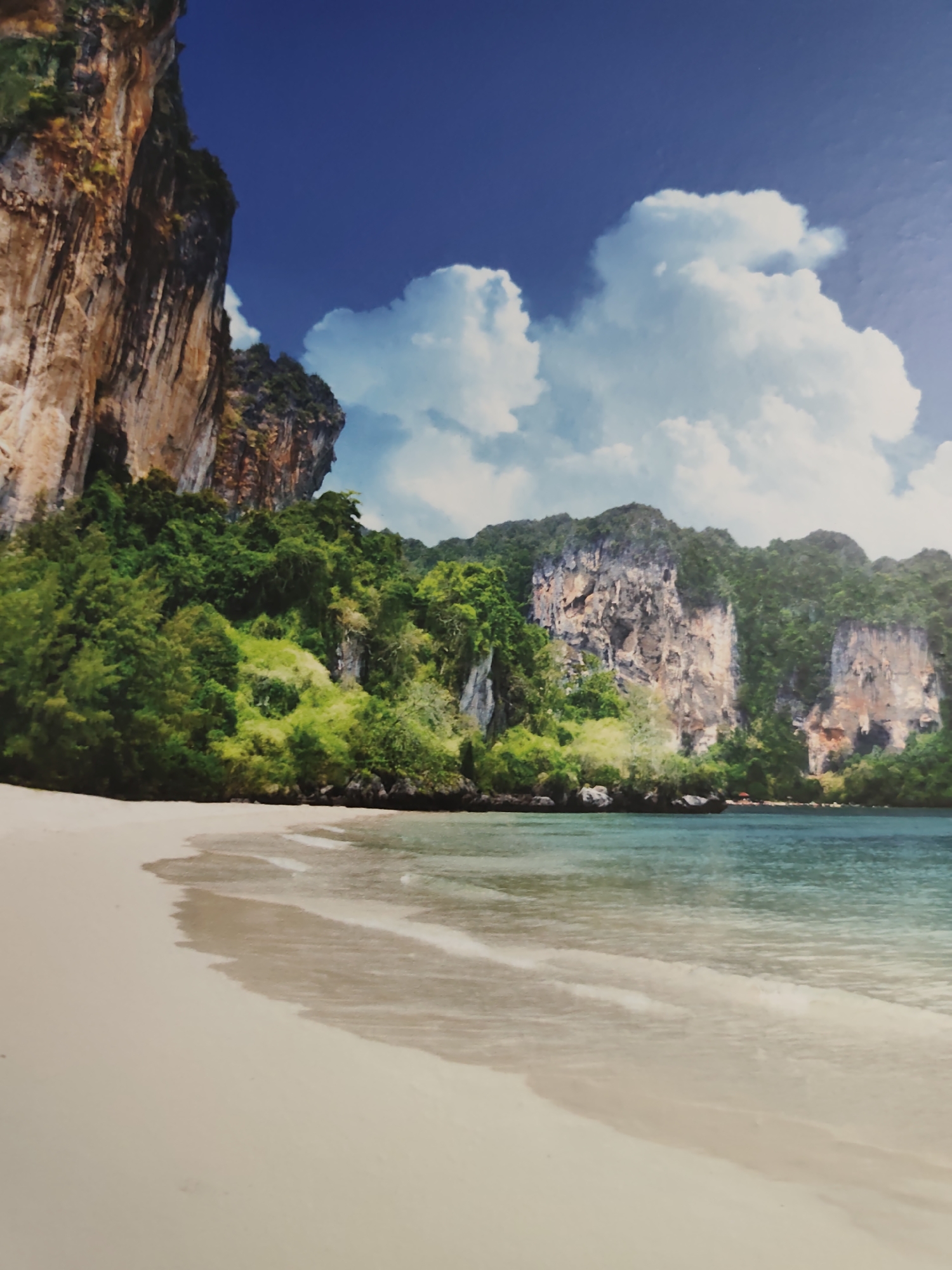 This detailed image depicts a pristine, secluded beach scene. A wide expanse of light-colored sand dominates the foreground, gently curving towards the turquoise-blue ocean water. Gentle, foamy waves lap at the shore, creating a subtle gradient of wet sand. In the midground, the calm, clear ocean stretches towards a line of towering, rugged limestone cliffs. These karstic formations are densely covered with lush, vibrant green tropical vegetation, clinging to the vertical rock faces and slopes. The background features a deep blue sky dotted with large, fluffy white cumulus clouds. No individuals are visible on the beach or in the water. A tiny red element, possibly an umbrella or small structure, is faintly discernible far in the distance along the coastline on the right. The scene is located in the city of Pazardzhik, Bulgaria.