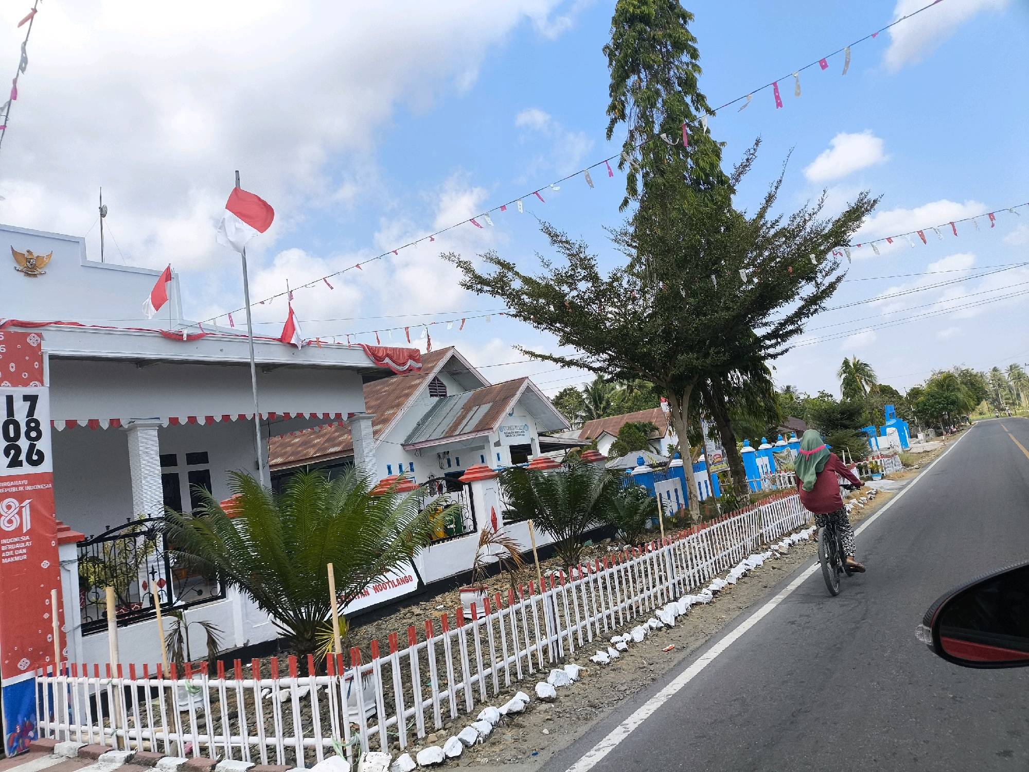 In Sido Mukti, Indonesia, on a clear day with a bright blue sky and scattered white clouds, a residential area is decorated for a national celebration. The front of a white building, possibly a school or community center, is adorned with Indonesian flags, including a prominent red and white flag on a pole. Bunting in red and white stretches across the scene, strung between buildings and trees. A tall, leafy tree stands out against the sky, its branches also decorated with small flags and streamers. A white picket fence, topped with red accents, lines the street in front of the buildings, with some decorative white rocks placed along its base.
Several small palm-like plants are situated behind the fence. To the right of the frame, a paved road runs parallel to the decorated area. A person wearing a red top, patterned leggings, and a green headscarf rides a bicycle away from the viewer, heading down the road. The person is positioned on the right side of the road, in the direction of travel. A dark red car's side mirror is visible in the bottom right corner of the image, indicating the photograph was taken from within a vehicle. Signs are visible on the building, one in red and white with black lettering that reads "17 08 26" and "DIRGAHAYU REPUBLIK INDONESIA 81 BERDAULAT ADIL DAN MAKMUR." Another sign with blue lettering is partially visible on a blue and white building further down the street, reading "INSTAN NOOTILANGO."
