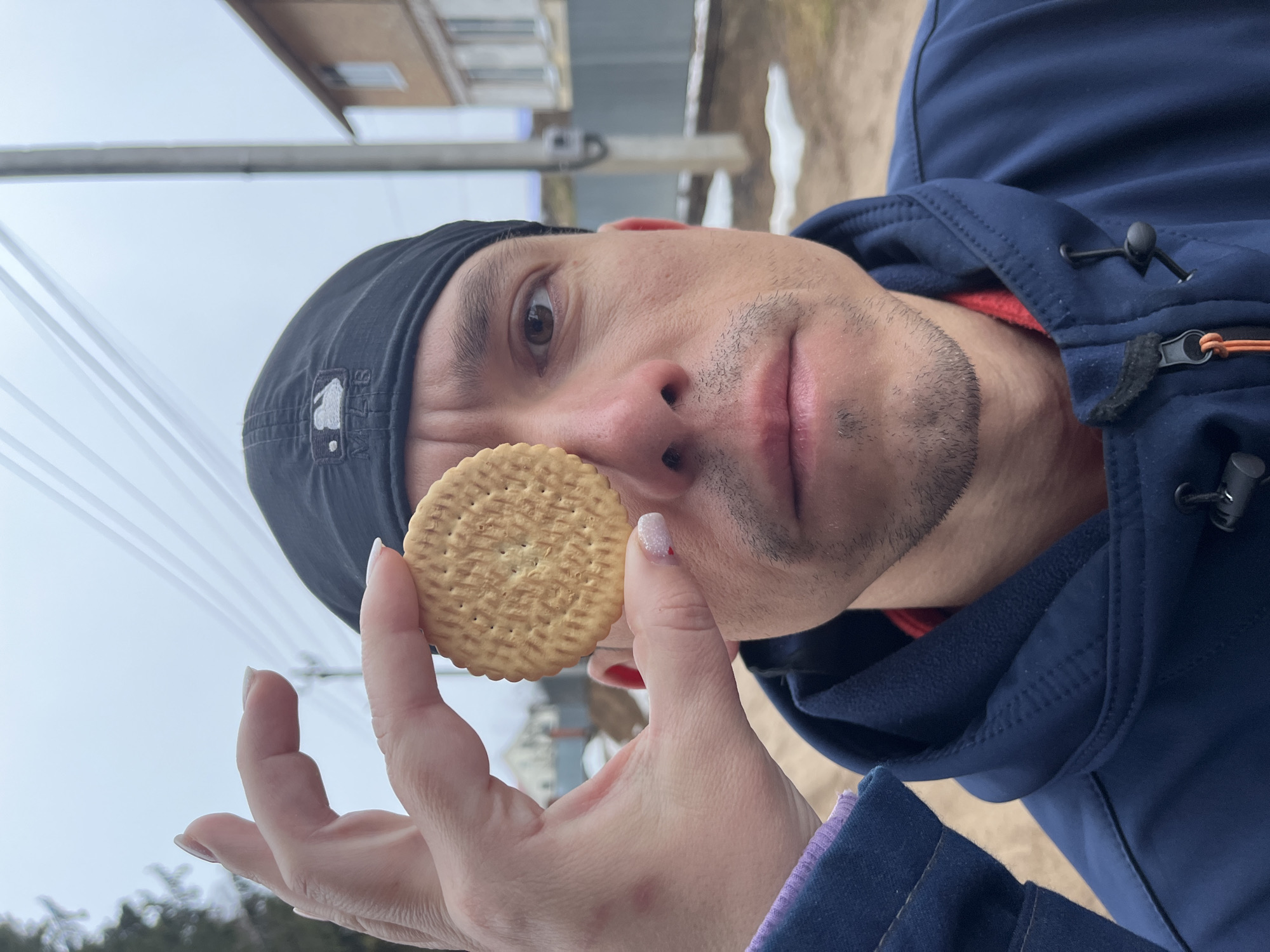 A man wearing a black cap and blue hooded jacket holds a round biscuit near his face. He has short dark hair and stubble. The biscuit is held by a hand with painted nails. In the background, there is a building and snowy ground, suggesting a location in the city of Chaikovsky, Russia. Overhead power lines are visible in the background.