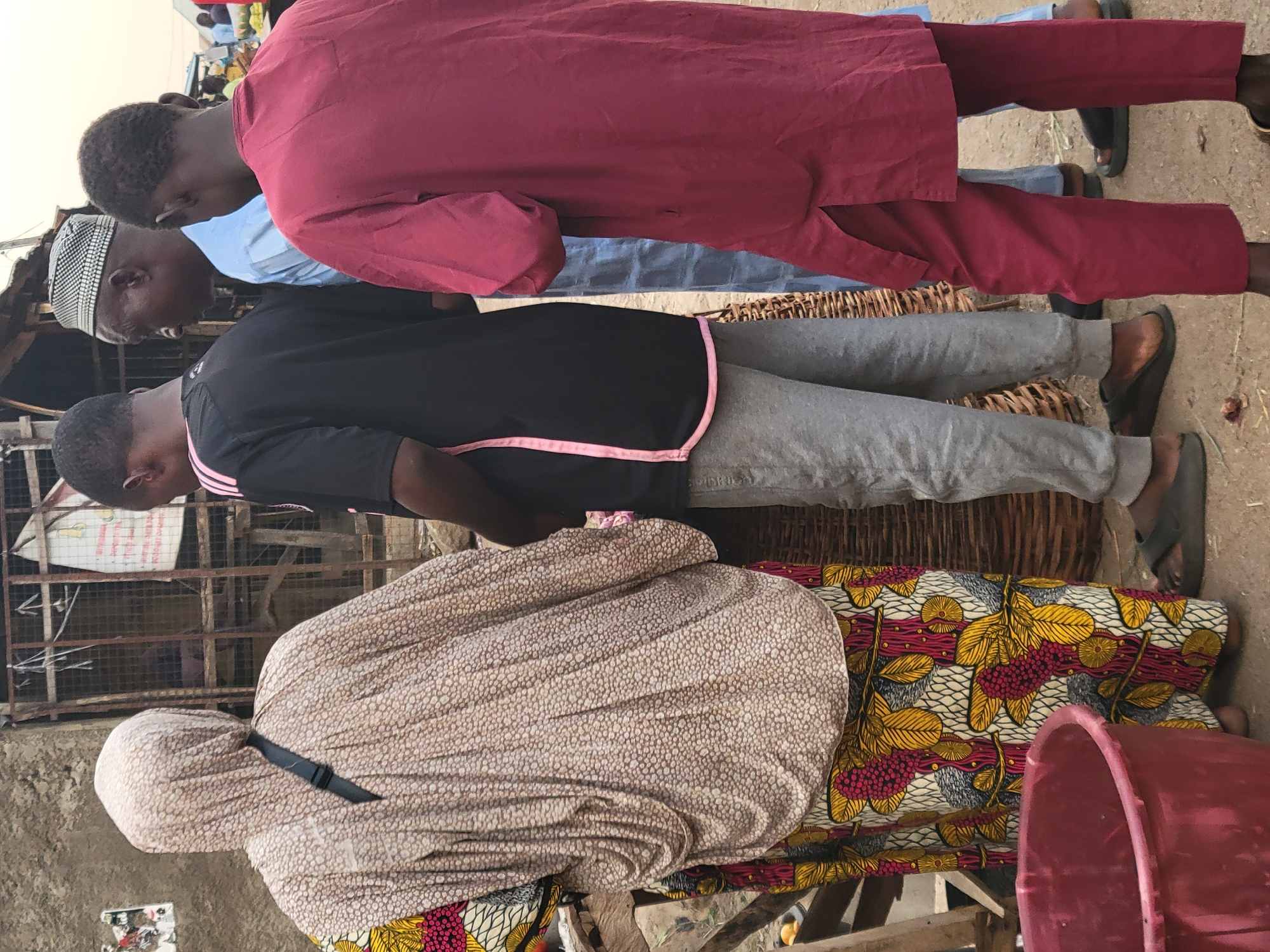 Four individuals stand in a market setting in Abuja, Nigeria. The person closest to the foreground is wearing a patterned hijab and a colorful printed fabric. Behind them, a person in a black t-shirt with a pink stripe and grey pants stands next to a woven basket. Behind them is a man wearing a traditional Muslim cap and a light blue shirt. Further back, a person in a red robe stands. The market environment is characterized by a mix of constructed walls, corrugated metal, and cluttered surroundings, indicating a bustling marketplace setting. A red plastic bucket sits near the woman in the foreground.