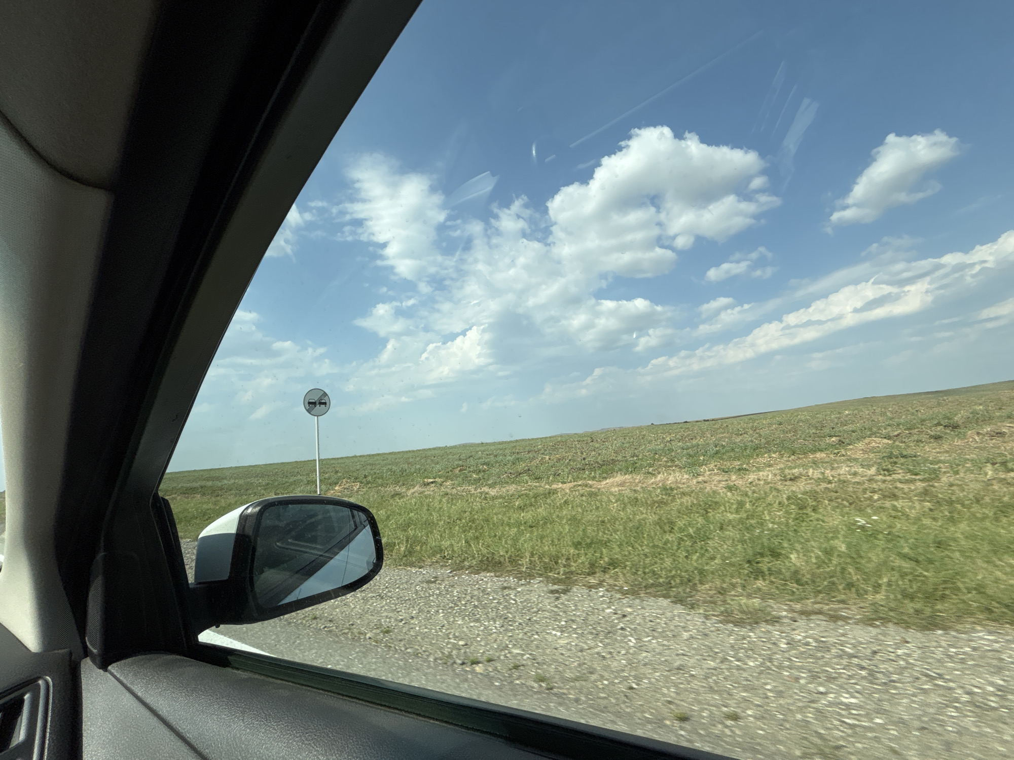 The image shows a view from the interior of a car, looking out the passenger-side window. The window frame and a portion of the car's interior paneling are visible in the foreground. Outside the window, a grassy field slopes gently upwards towards the horizon under a blue sky with scattered white clouds. A white road sign with a red circle and a diagonal red line through two car symbols is visible to the left of the field. The sign indicates a prohibition on overtaking. The car is on a gravel road or shoulder. The scene appears to be in a rural or semi-rural area, consistent with the location of Nagornoye, Russia, known for its open landscapes. There are no people visible in the image.