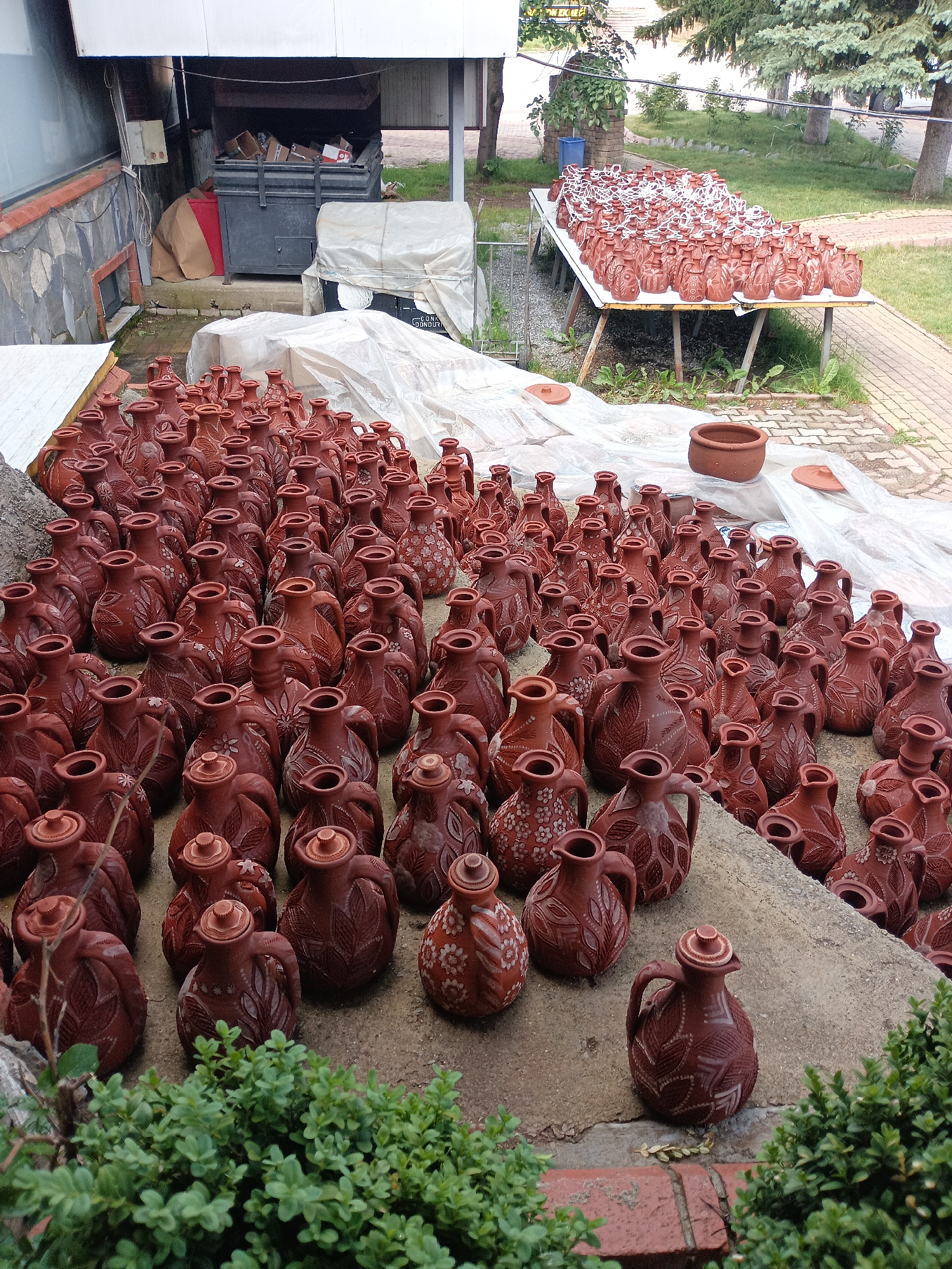 An outdoor scene in Seydişehir, Turkey, showcases a pottery workshop or storage area. Numerous clay jugs, many adorned with intricate carved patterns of leaves and flowers, are arranged in dense rows on a concrete surface. Some jugs are covered with translucent plastic sheeting. In the background, a large metal dumpster filled with cardboard boxes sits near a stone-walled building. Beyond this, a long white table is covered with more clay jugs, this time with white string tied around their necks. A single, larger brown pot sits on the plastic sheeting, and other smaller clay lids are scattered nearby. The environment is an outdoor yard with grass and trees visible, suggesting a warm, possibly sunny day.