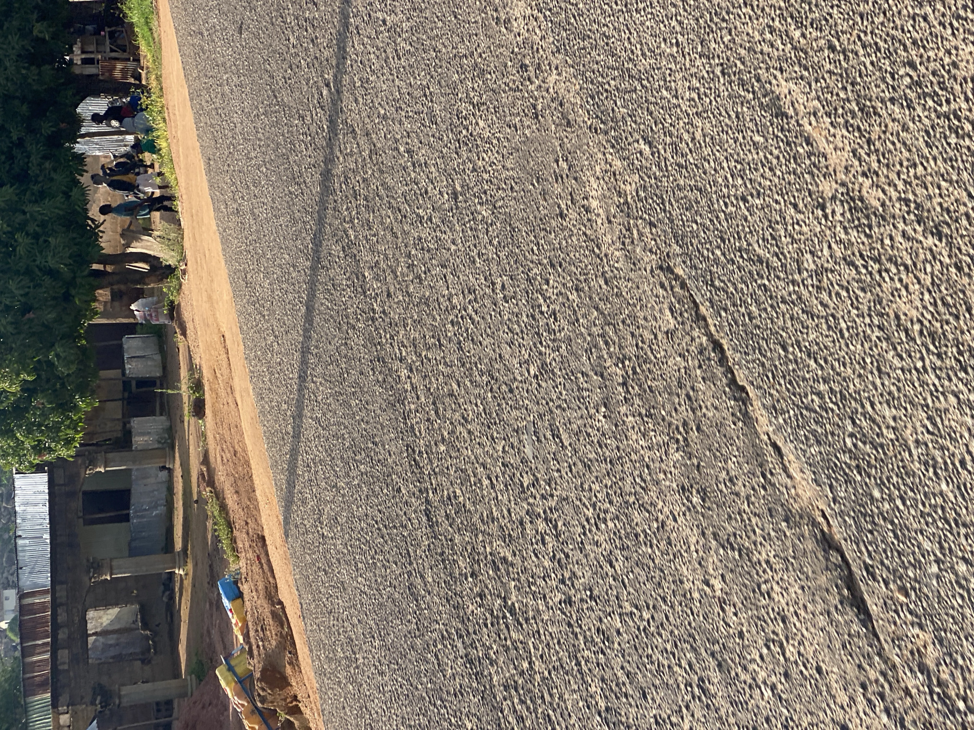 In Jos, Nigeria, a paved road with visible cracks runs diagonally across the frame. To the left, a dirt embankment slopes upwards, littered with colorful plastic bags and what appears to be construction debris. Beyond the embankment, the ruins of a building with stone columns and a corrugated metal roof are visible. To the right of the embankment, several people are gathered under the shade of trees. Some are standing, while others are seated. They appear to be engaged in conversation. The overall scene suggests a rural or semi-urban environment with signs of both development and decay.