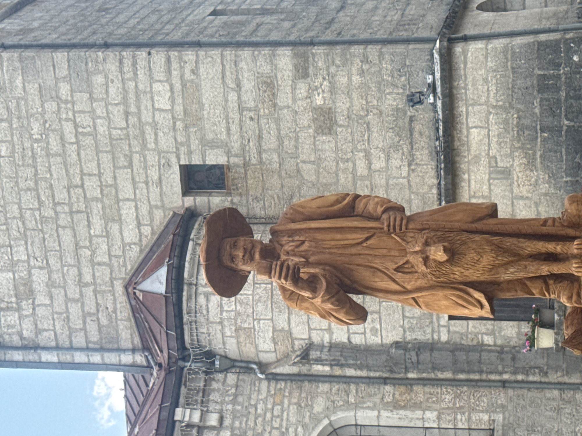 A wooden statue of a man wearing a wide-brimmed hat and a cape stands in front of a stone building. The man is holding a small object in his right hand and has a wolf statue at his side. The stone building appears to be a church or historical structure, with a peaked roof and arched windows visible. The sky above is partly cloudy. This statue is located in the city of Grande-Rivière Château, France.