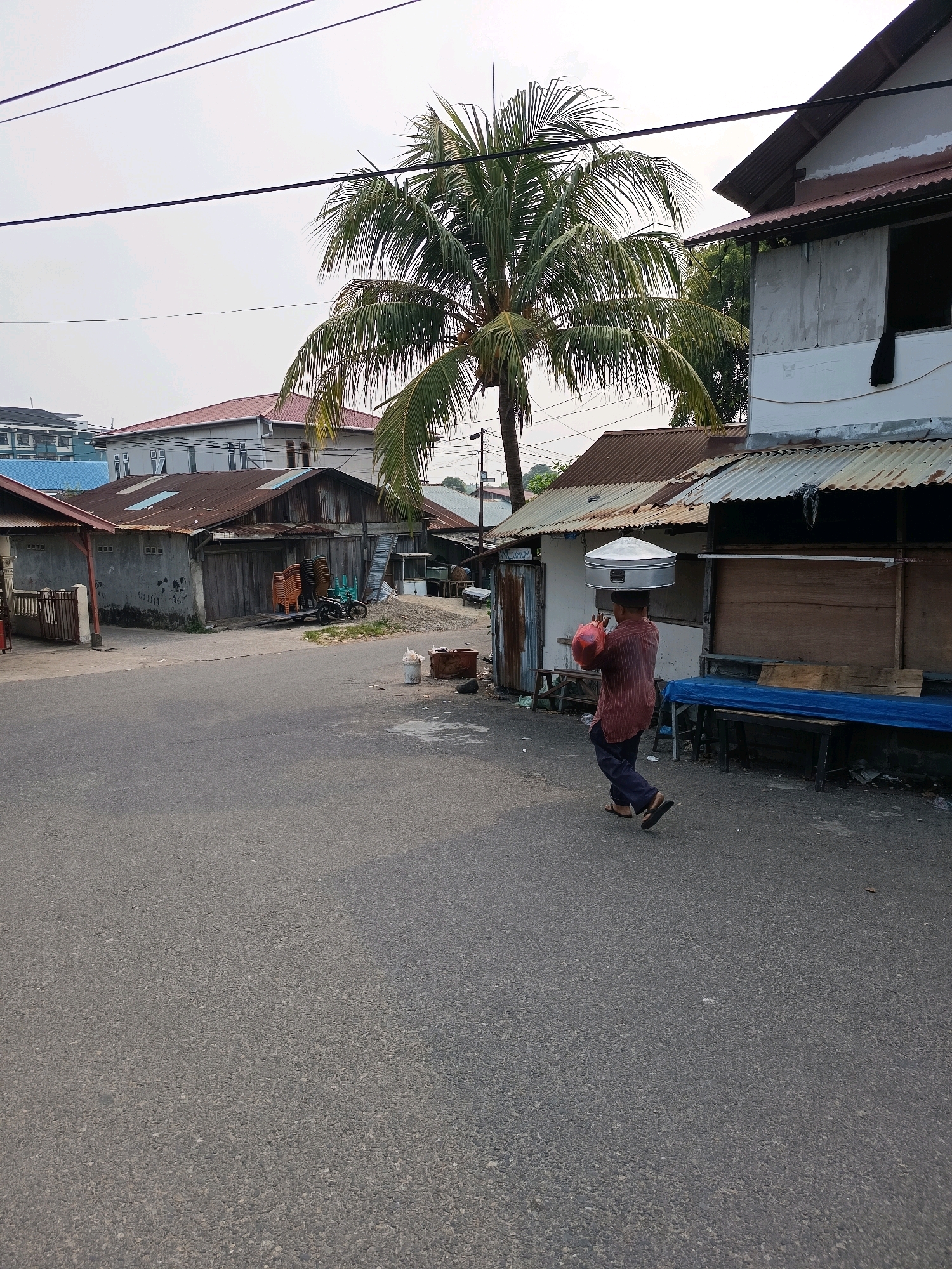 The image depicts a street scene in Padang, Indonesia. A person is walking on the asphalt road, carrying a large silver pot on their head and a red plastic bag in their right hand. The person is wearing a striped maroon shirt and dark blue pants, with sandals. Behind the person, there are several buildings with corrugated metal roofs. Some of these buildings appear to be shops or stalls with wooden shutters. A palm tree with green fronds is visible in the background, with power lines running across the sky. The environment appears to be a mix of residential and commercial buildings. The sky is overcast, suggesting a cloudy day.
