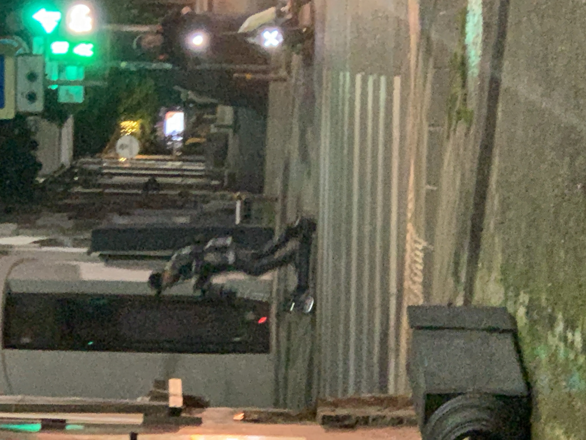 The image depicts an urban street scene at night or dusk, characterized by artificial lighting and a damp ground surface, suggesting recent rain. The camera perspective is slightly elevated, looking down onto the street.
In the mid-ground, a person wearing dark clothing is visible, appearing to be on a skateboard or similar wheeled device at a crosswalk. Their posture suggests they are either crouching, sitting, or preparing to move. The crosswalk itself features white stripes, and faint, illegible text appears to be embossed on its surface.
To the left of the scene, a traffic light pole displays two illuminated green lights: one is a clear left-turn arrow, and another is a solid green light. Below these, a rectangular signal box is mounted. Further in the background, other blurry figures and vehicles are discernible, including what appears to be a vehicle with bright lights (headlights or taillights) on the far side of the intersection. Another bright, "X"-shaped light, possibly from a scooter or similar personal electric vehicle, is visible in the upper background.
On the far right, green foliage from trees or bushes is visible. The foreground on the bottom right features a dark, rectangular street barrier or planter, with what looks like a wheel or tire partially visible. Buildings or concrete structures line both sides of the street. No discernible legible text is visible other than the faint, unreadable text on the crosswalk.