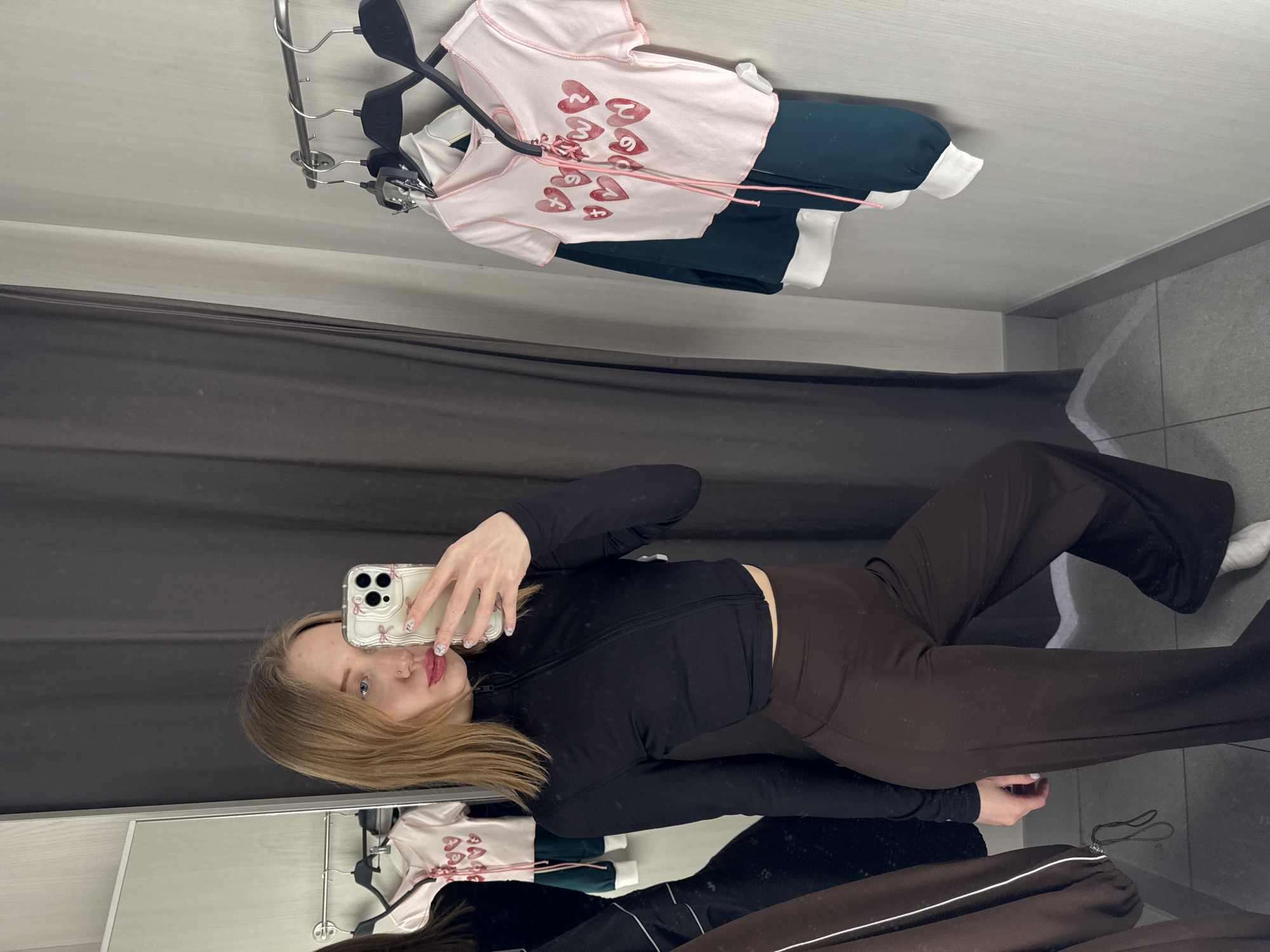 A young adult female is depicted in a retail store fitting room in the city of Ovoshchnoy, Russia. She is taking a mirror selfie, holding a smartphone with a white, pink-bow-decorated case in her right hand. Her nails are manicured with white polish and pink designs. She has shoulder-length light brown hair, blue eyes, a small silver nose stud, and pink lipstick. She is wearing a black long-sleeved cropped top with a partial zipper and dark brown wide-leg pants, with white footwear partially visible.
The fitting room features a large vertical mirror on the left, reflecting her image and a dark brown garment. A dark gray curtain is visible on the left and behind the reflection. The right wall is light gray and textured, fitted with a silver metal clothes rod holding several black hangers. Garments hanging on the rod include a light pink short-sleeved top with red heart graphics and pink lacing, layered over a dark green long-sleeved top with white cuffs and hem. The floor is covered with gray tiles, and a dark gray baseboard lines the walls.