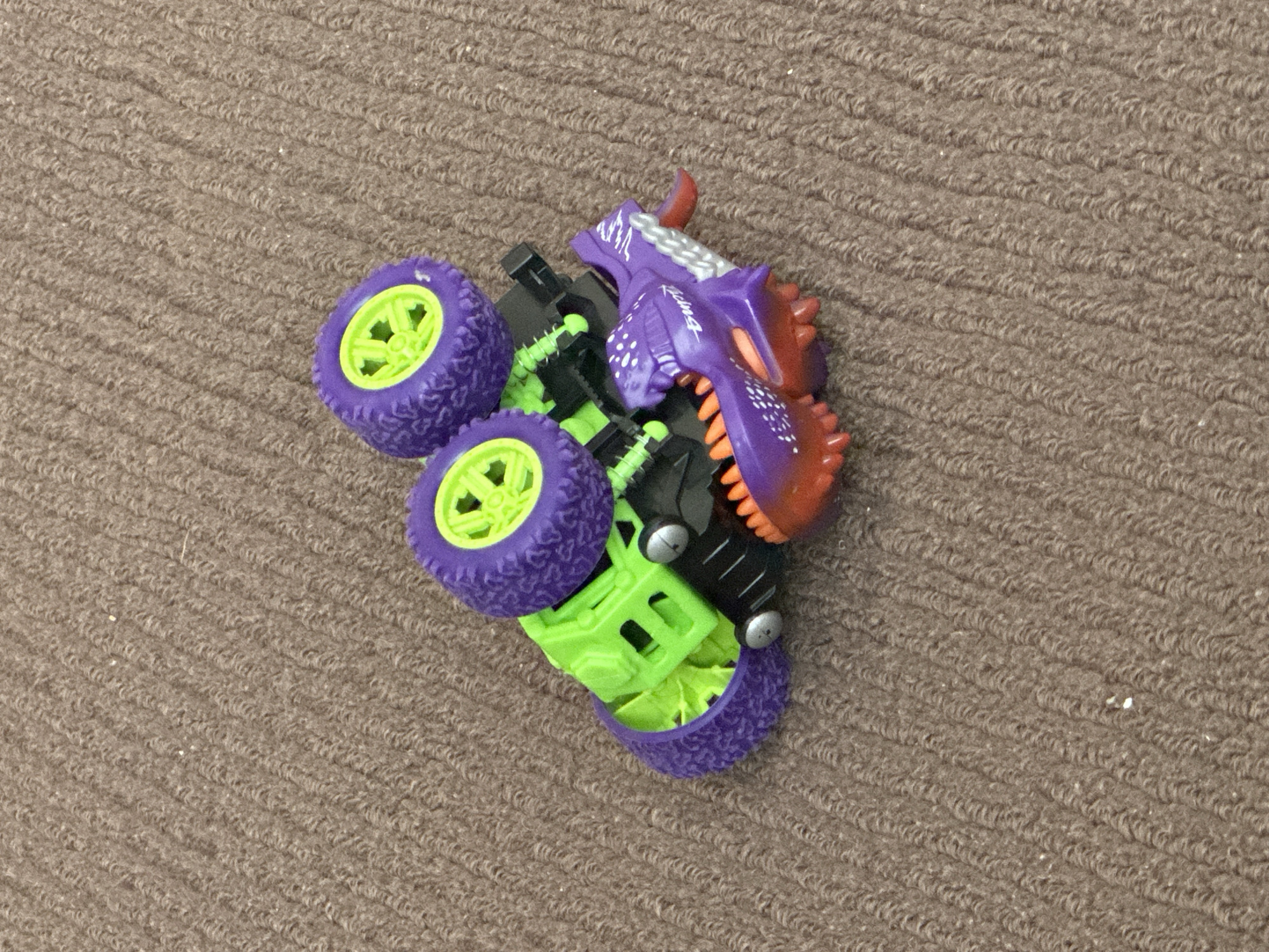 A close-up, high-angle shot shows a toy monster truck on a brown, looped carpet. The truck is predominantly lime green and purple with a purple, dragon-like head attached to the front. The dragon head is open-mouthed, revealing orange teeth and a red tongue. The truck has four wheels; two are clearly visible, one purple with a lime green rim, and another partially visible purple wheel. The truck is flipped onto its side, with the green chassis and black suspension components exposed. The carpet texture is very detailed, with visible individual loops. The image appears to be taken indoors, possibly in a home environment. There are no people visible in the image. The location in the city of Trieste, Italy, is not discernible from the provided image.