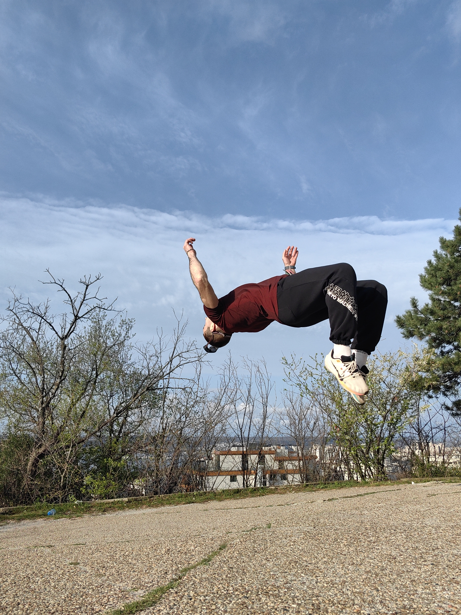 A person is performing a backflip outdoors. The individual wears a maroon t-shirt, black sweatpants with white lettering on the right leg, white socks, and sneakers. They are in mid-air against a backdrop of a cloudy sky, trees, and buildings. The ground appears to be a gravel surface. The location is likely in Varna, Bulgaria, given the specified information. The photograph suggests an urban environment near a park or open space, possibly on a hill overlooking the city.