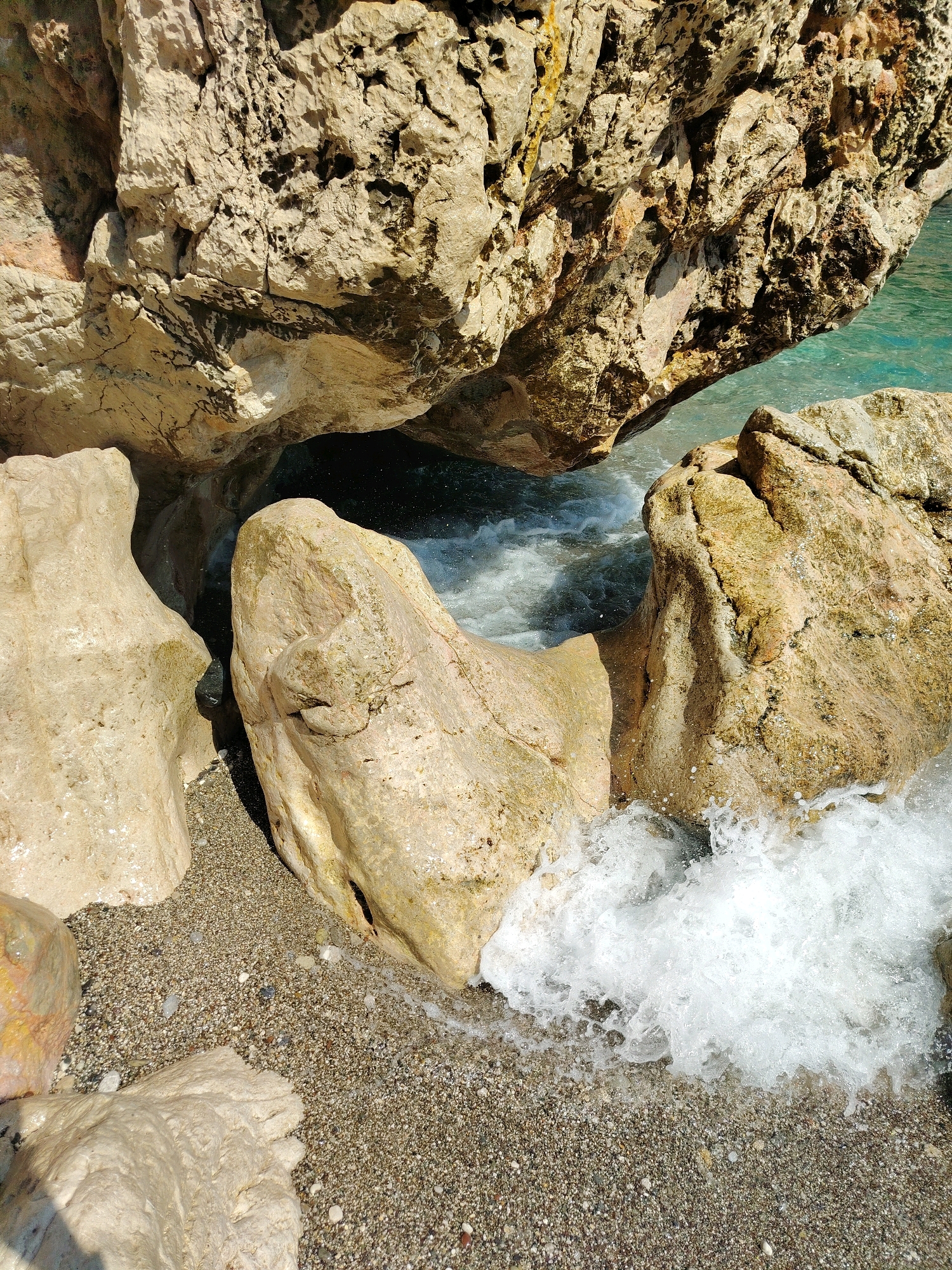 The image depicts a coastal scene with large, weathered rocks and the sea in the city of Kemer, Turkey. The rocks are predominantly light brown and beige, with textured surfaces and visible cracks and crevices. Some rocks are large and imposing, forming natural formations, while others are smaller and scattered on a pebble beach. The sea is a vibrant turquoise color, with clear water visible around the rocks. Waves are crashing against the rocks, creating white foam and splashes. The pebble beach is composed of small, rounded stones in various shades of gray and brown. The lighting suggests it is daytime, with sunlight illuminating the scene and casting shadows. There are no people or animals visible in the image. The overall impression is a natural, rugged coastal landscape.