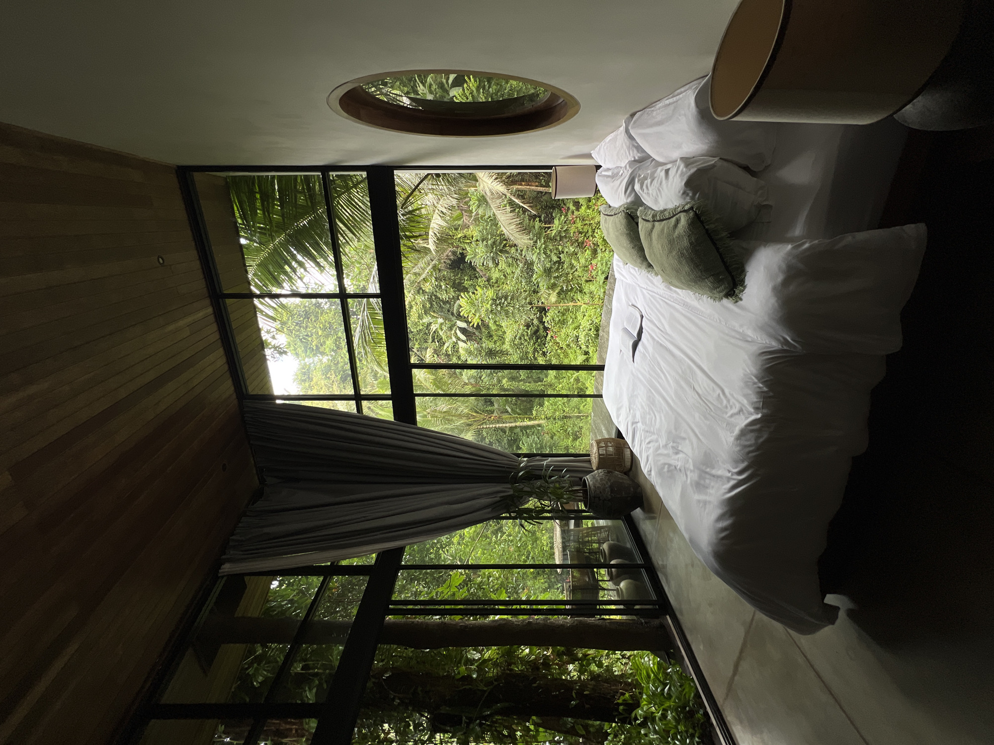 The image depicts a bedroom in a modern dwelling, possibly a villa, with extensive floor-to-ceiling windows overlooking a dense, verdant forest. The room is minimalist and chic, featuring a large bed covered with crisp white linens and adorned with two textured green throw pillows. A book lies open on the bed, suggesting recent use. The flooring appears to be polished concrete, and the ceiling is paneled with dark wood, creating a sophisticated contrast. A large, grey curtain hangs on the left side of the room, partially obscuring the view. On the right wall, a circular, wooden-framed mirror reflects the lush greenery outside. A bedside lamp with a neutral shade is visible on a dark base. The overall impression is one of serene luxury, seamlessly integrated with the natural environment. The location is identified as being in the Ubud District, Indonesia, which is renowned for its lush rainforests, rice paddies, and vibrant cultural scene, aligning with the visual elements of the image. There are no people present in the scene.