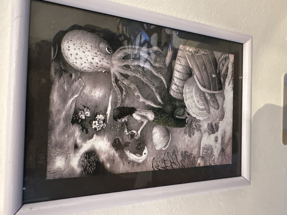 The image displays a framed, predominantly grayscale artwork depicting an underwater scene, mounted on a light-colored wall in El Pinto, Spain. The illustration features a mermaid centrally positioned, possessing dark hair and a scaly, dark green tail. She holds a small, light-colored, duck-like creature in her hands, with small bubbles visible around it. To the upper right, a large octopus extends multiple tentacles across the scene.
The underwater environment includes a detailed seafloor with various marine elements. Prominent objects on the seafloor comprise large seashells, including a ribbed oyster shell in the lower right, and broken wooden structures resembling barrels or boat fragments. Marine life observed beyond the main subjects includes a long, light-colored fish near the mermaid's left, two striped fish in the upper-center, and a smaller, elongated fish in the mid-right. A starfish is visible on the seafloor in the lower-left. Coral-like formations and aquatic plants are distributed throughout the scene, with patterns suggestive of light filtering through water present in the upper-left quadrant. The artwork is enclosed within a simple white frame, and reflections are discernible on its glass surface.