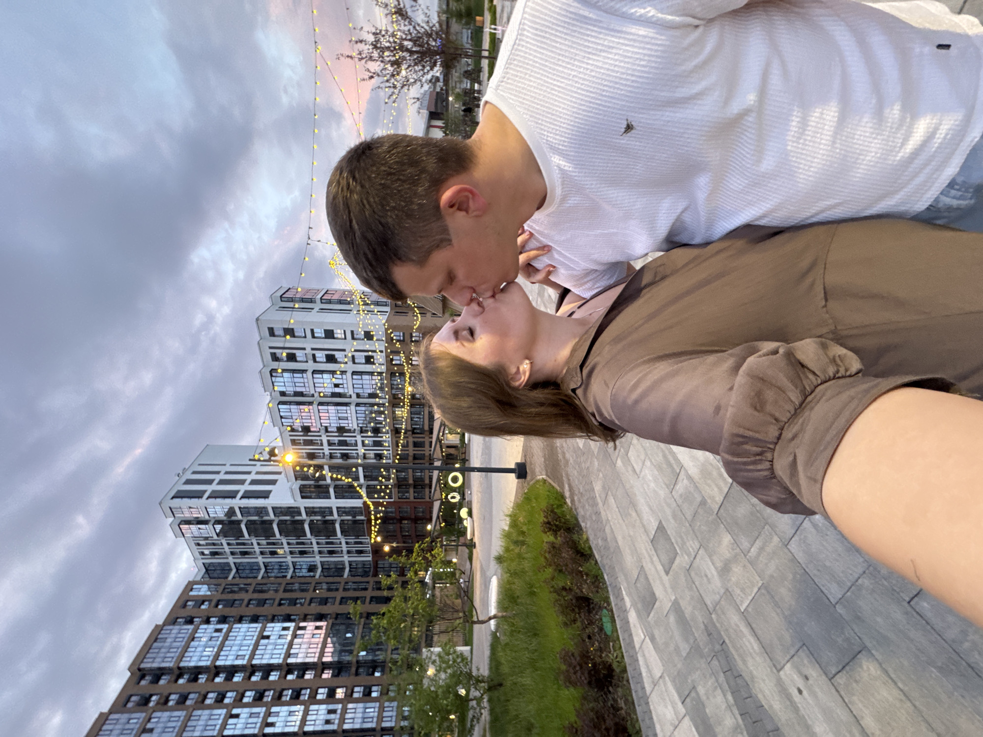 A couple is kissing in a paved area lined with green grass and trees. Multiple tall buildings are visible in the background, decorated with string lights. The sky is cloudy with a hint of color from the setting sun. The man is wearing a white shirt, and the woman is wearing a brown shirt. The image may be taken in Perm, Russia.