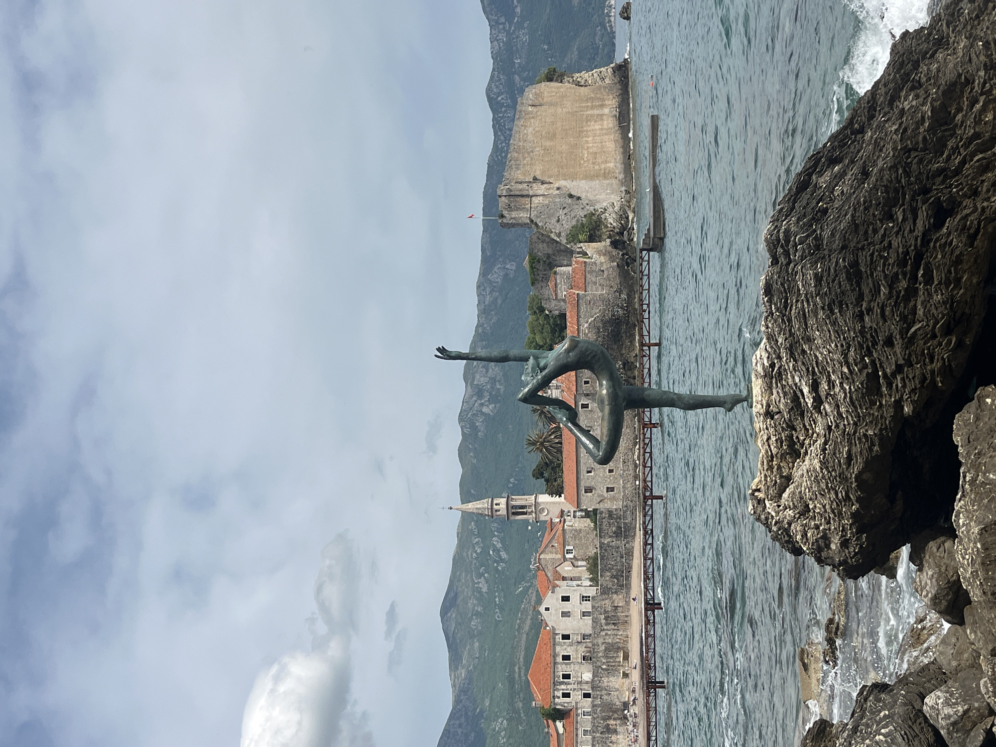 The image shows a bronze statue of a female gymnast in Budva, Montenegro, standing on rocks near the sea. The old town of Budva, with its characteristic red-tiled roofs and church tower, is visible in the background, along with the fortress on the peninsula. The sky is overcast. The statue is slender with an arching back and one leg extended skyward. The sea is choppy, and the rocks are dark and rugged.