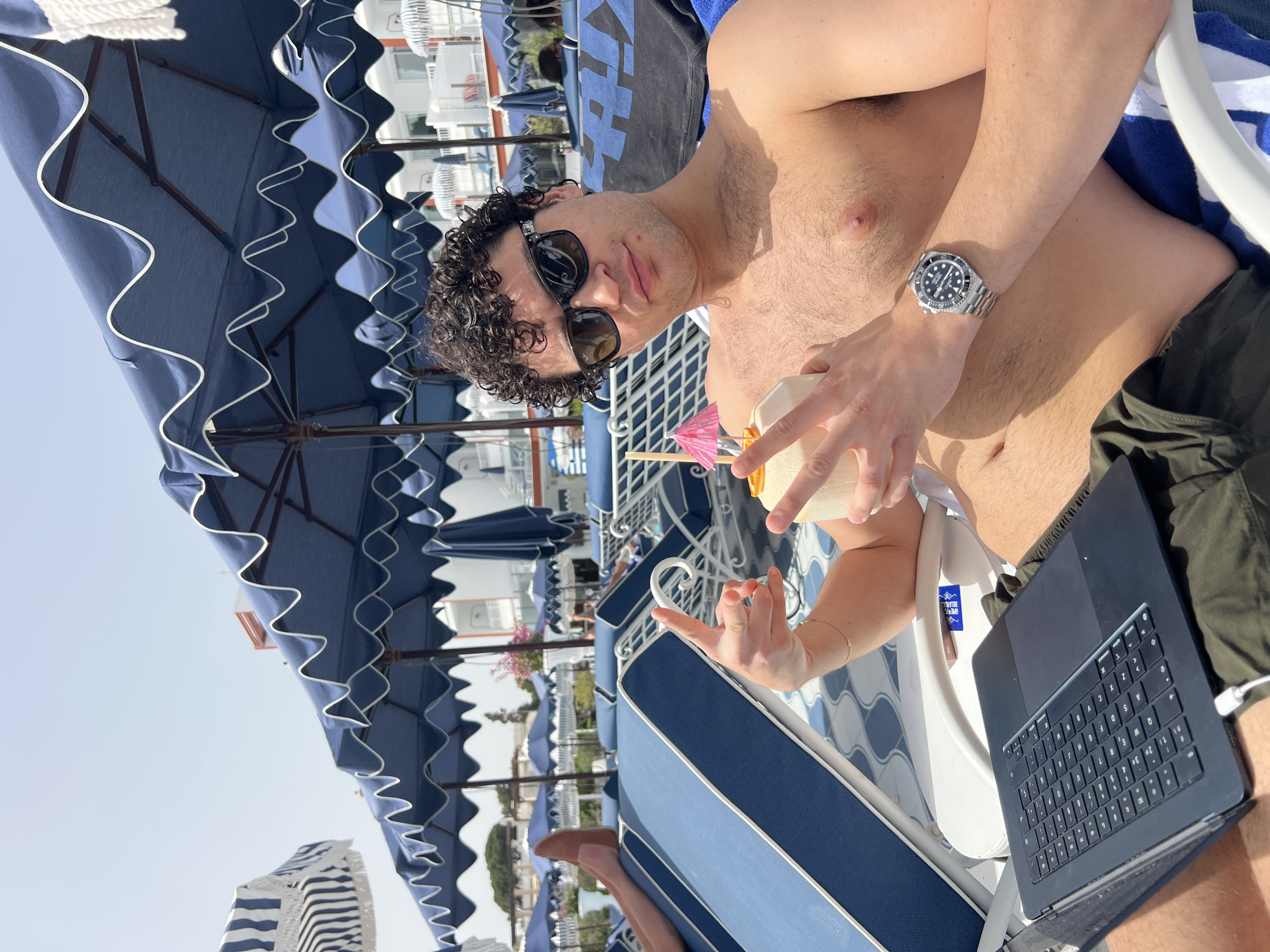 The image shows a man relaxing on a blue-and-white striped chaise lounge at a resort. He is shirtless, wearing dark shorts, sunglasses, and a wristwatch. He is holding a coconut drink with a small umbrella and has a laptop on his lap. The background includes blue-and-white striped parasols, resort buildings, and other chaise lounges. Based on the architectural style of the buildings and the distinctive parasols, the location is likely a resort in Santa Eulària des Riu, Spain.