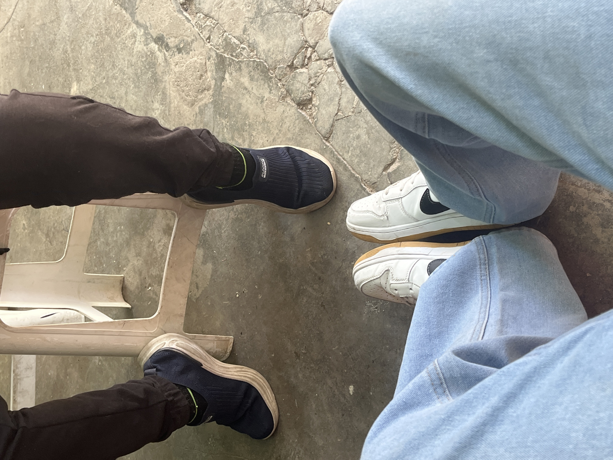 The image, taken in Juan Griego, Venezuela, provides a low-angle, downward perspective focusing on the lower legs and feet of three individuals on a concrete floor. In the immediate foreground, a person is seated or standing, wearing light blue denim jeans and white low-top sneakers with brown/gum soles and black accents, possibly a Nike swoosh, on the inner sides. Two other individuals are positioned in the mid-ground, observed from the foreground perspective. The person on the left wears dark brown or black athletic pants and a dark blue slip-on athletic shoe with a light beige sole and black socks with a visible lime green top edge; their left foot is resting against the leg of a white, somewhat dirty, plastic chair. The person on the right wears similar dark brown or black athletic pants and a matching dark blue slip-on athletic shoe with a light beige sole and similar socks. The dull gray concrete floor exhibits numerous fine cracks and general wear, with areas of varying discoloration.