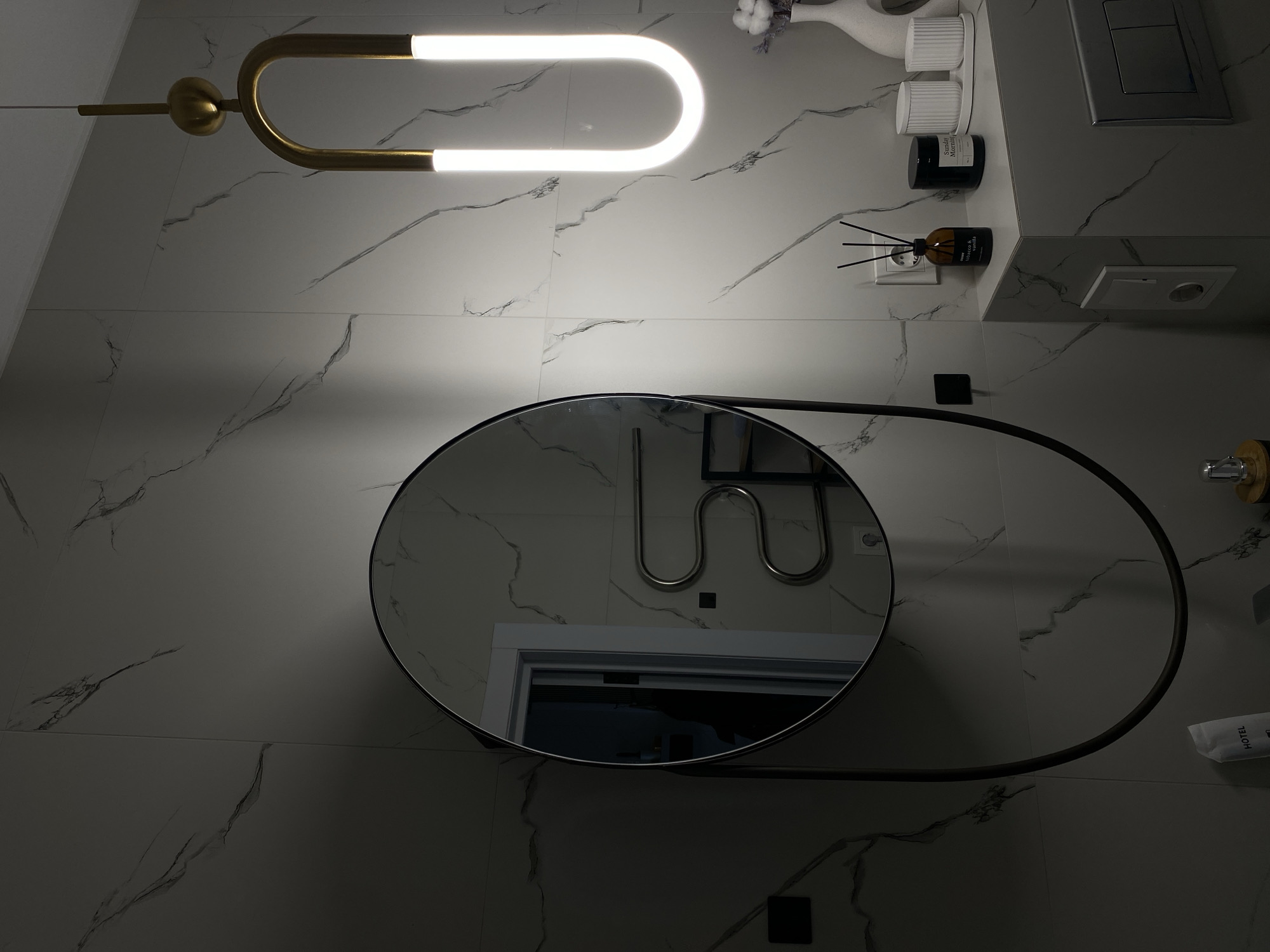 This image captures a modern, well-lit bathroom interior, likely within a hotel, characterized by its clean aesthetic and marble-patterned white wall tiles. The perspective is tilted, looking down into the room.
A prominent feature is a unique, gold-finished light fixture in the upper left, boasting a bright, U-shaped LED element that casts a significant glow. Below this, a large, round mirror with a sleek dark frame dominates the central part of the wall. Its reflection reveals an adjacent area, including a white door frame and a silver, serpentine-shaped towel warmer or heater.
On a counter or shelf to the right, several items are neatly arranged: a white vase holding decorative branches (possibly cotton), two ribbed white cups or containers, and a dark jar with partially visible text "Sunday Morning". Further along the shelf, a brown glass diffuser with black reeds is seen, plugged into a wall socket. Its label features the words "tobacco & vanilla" and "ROOM". A silver flush plate is mounted on the wall above the diffuser. In the bottom right corner, a white packet labeled "HOTEL" indicates the location is a lodging establishment.
The scene is static, with no activity taking place. The artificial lighting suggests an indoor setting, precluding any inference about the time of day or outdoor weather. The overall impression is one of contemporary design and functional elegance, consistent with a modern hotel in Kazan, Russia.