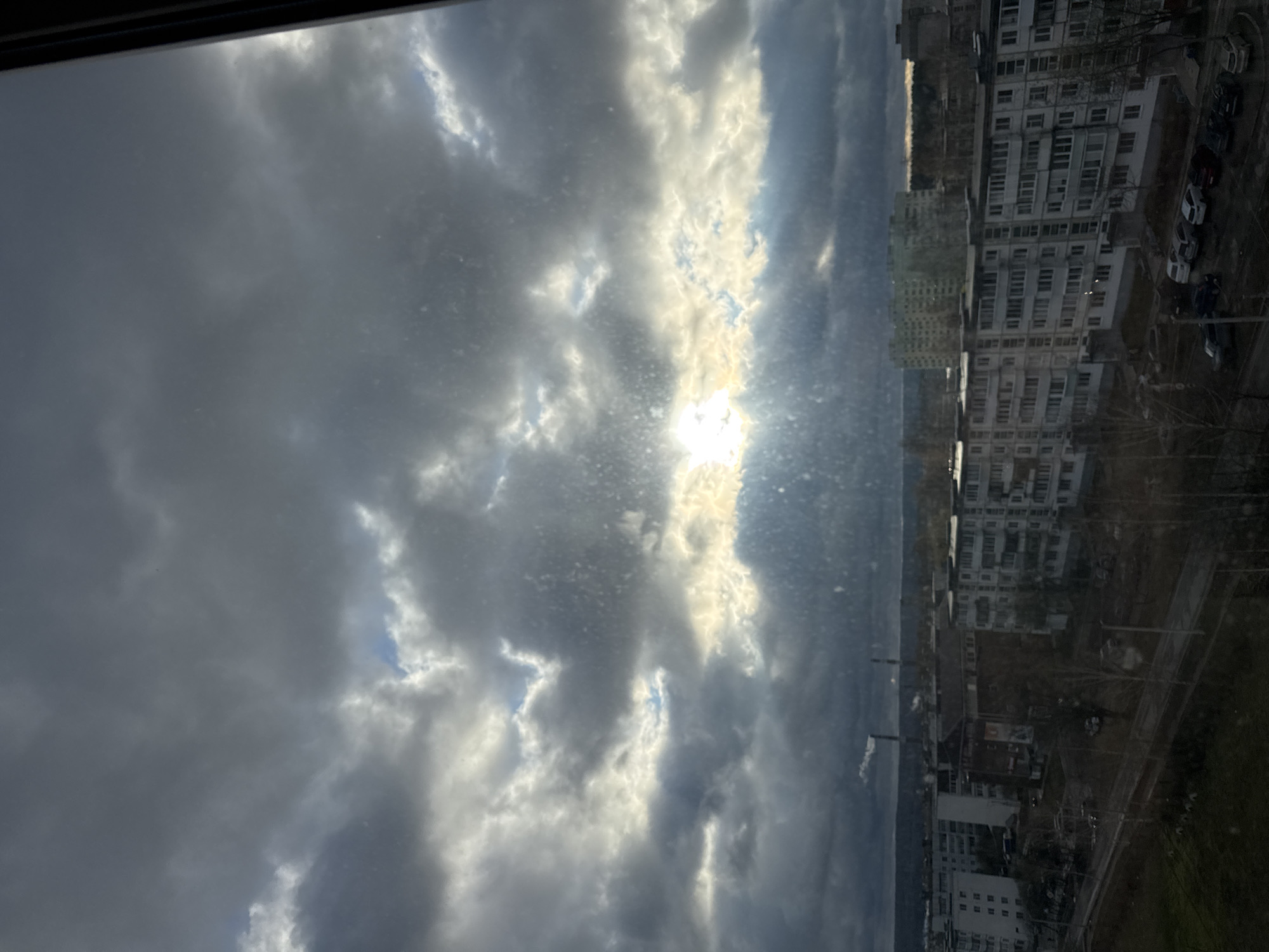 The image, taken from an elevated perspective through a window, captures an urban landscape under a cloudy sky in Chaikovsky, Russia. The sky dominates the upper portion of the frame, filled with dense, textured grey and white clouds. A bright, powerful sun breaks through a gap in the clouds in the upper-middle right, casting strong light and creating dramatic contrast. Scattered patches of blue sky are visible between the cloud formations. Small specks and smudges are visible on the window surface through which the photograph was taken.
Below the horizon, an urban environment unfolds, primarily featuring residential buildings. On the right side, multiple multi-story apartment blocks, characteristic of post-Soviet architectural styles with their uniform facades and balconies, line the streets. Several cars are parked along the street adjacent to these buildings. In the hazy distance, particularly on the left side of the mid-ground, a tall industrial chimney is visible, emitting a light plume of smoke. The time of day appears to be daytime, likely late morning or afternoon, with the sun providing intense illumination through the cloud cover. No people are visible, and there is no discernible text within the image.