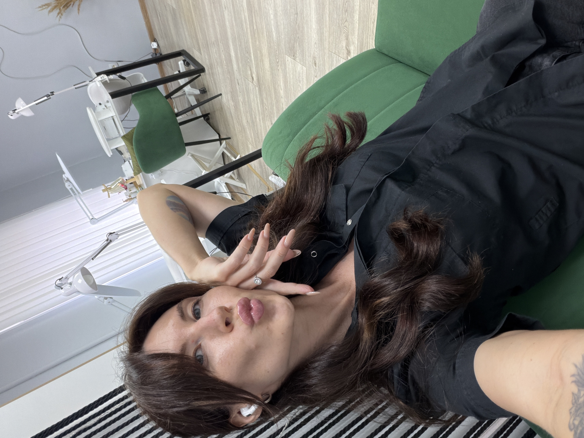 A woman is taking a selfie in a room that appears to be a nail salon. She is wearing a black button-up shirt and has dark brown hair. She is leaning her head against her hand and puckering her lips. There are nail lamps and a manicure table in the background, along with a green chair. The flooring is light wood. The environment suggests an indoor commercial space.