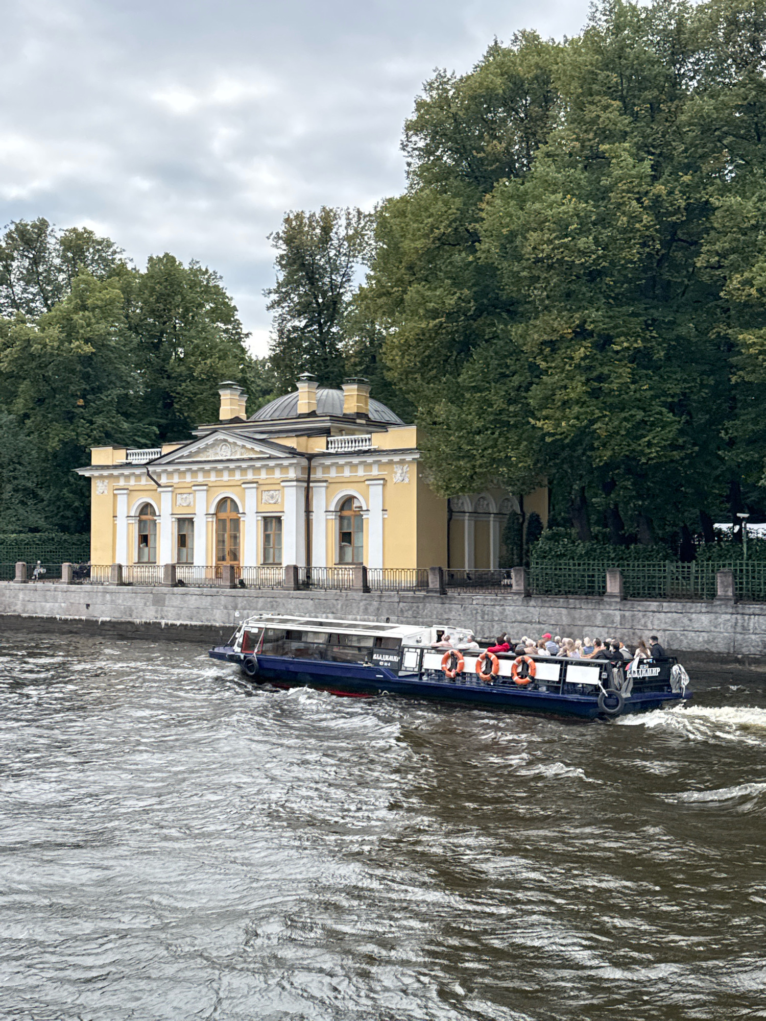 The image shows a tour boat with passengers on a body of water in front of a yellow neoclassical building and lush green trees. The building, with its white columns and arched windows, appears to be a pavilion or a small palace. The boat, with its white roof and blue hull, is moving from left to right, creating ripples in the water. The passengers are seated on the deck, and some are looking at the scenery. The trees in the background are dense and full, suggesting it is summer or early autumn. The sky is overcast with gray clouds. This scene is likely located in the Catherine Park in Pushkin, a suburb of Saint Petersburg, Russia, known for its palaces and gardens. The building is likely the Hermitage Pavilion.