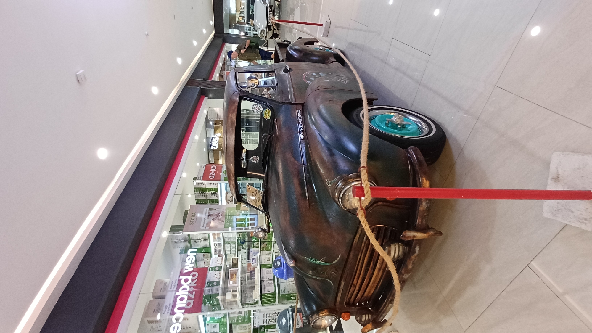 The image shows a vintage car on display inside a shopping mall. The car appears to be a heavily modified, rusted hot rod with turquoise-colored rims. It is surrounded by red poles connected by rope. Behind the car is a storefront with shelves filled with items. There is a man is partially visible in the distance inside the car. The location is Kuta, Indonesia.