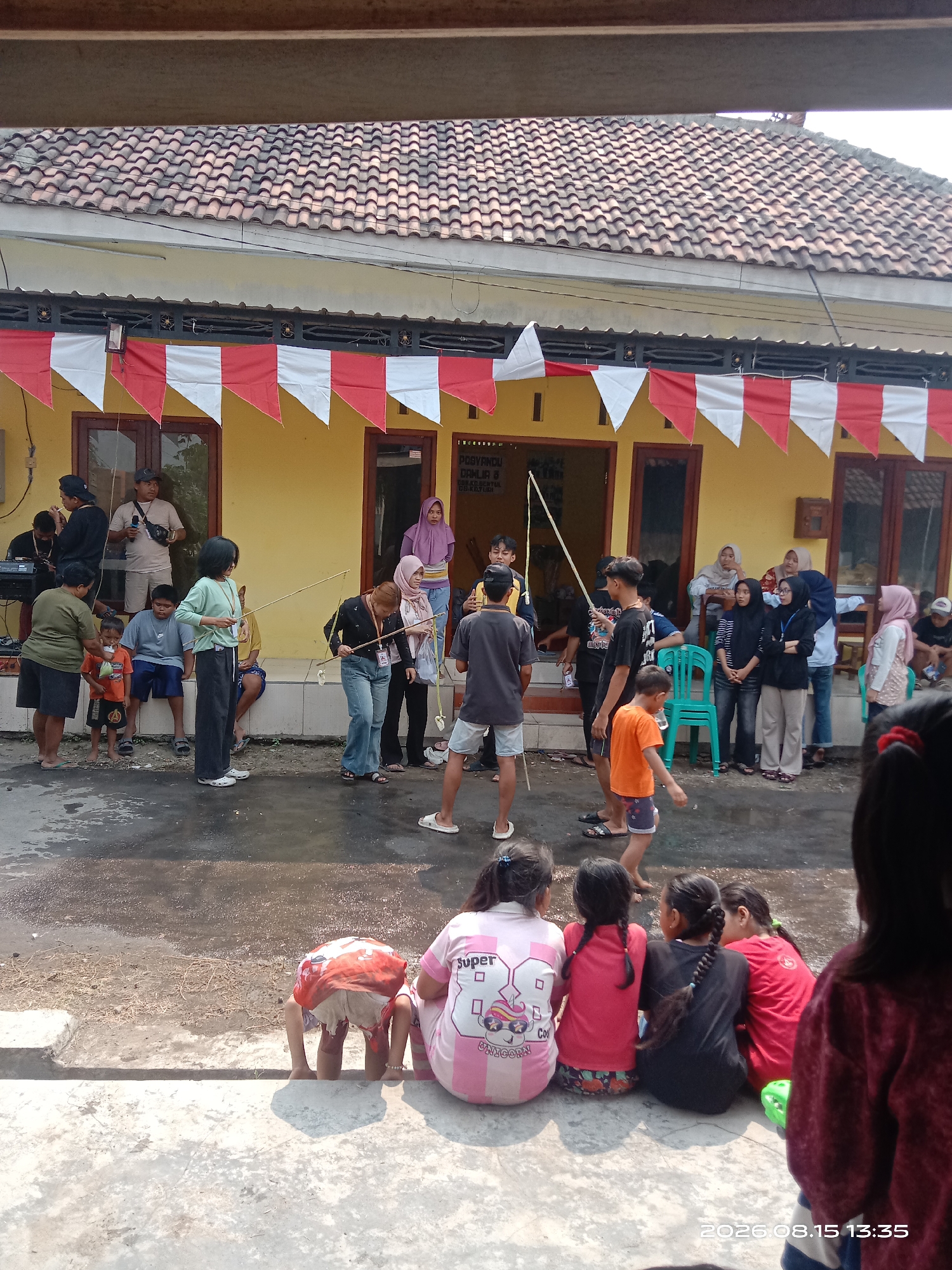 The image depicts an outdoor scene in Banyuarang, Indonesia, with a group of people gathered in front of a yellow building decorated with red and white triangular banners. The building has a tiled roof and several windows and doors. Several individuals are engaged in what appears to be a game or activity involving fishing rods with some sort of bait. Some people are standing and observing, while others are sitting on the ground or on a low wall. There are children present, some playing or watching the activity. The ground is paved with concrete and asphalt, with some wet patches indicating recent rain or water. The overall atmosphere suggests a community event or gathering.