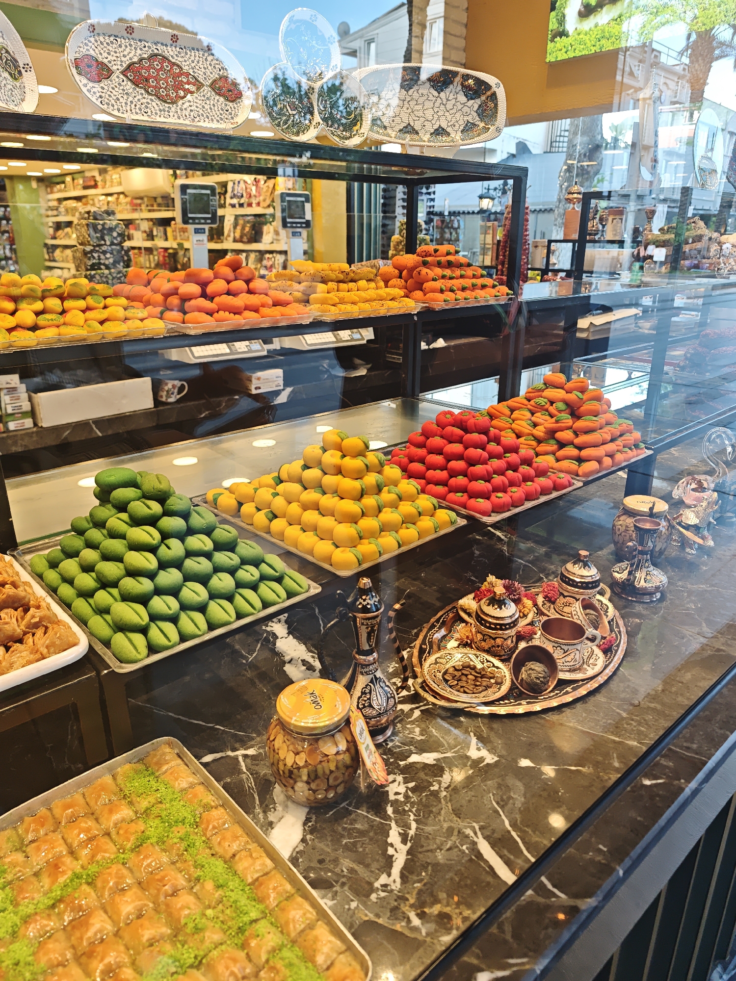 The image depicts a display of Turkish sweets in Kemer, Turkey. Assorted brightly-colored sweets, formed into spherical and oblong shapes, are stacked in pyramids on serving trays within a glass display case. On the lower level, a tray of baklava sprinkled with green pistachio nuts is visible. A jar of nuts in honey and a decorated Turkish coffee set on a tray are placed to the side, suggesting the products are part of a dessert offering. In the background, shelves stocked with products are visible through the display case, indicating the location is likely a shop or market. The overall setting suggests a commercial establishment specializing in confectionery.