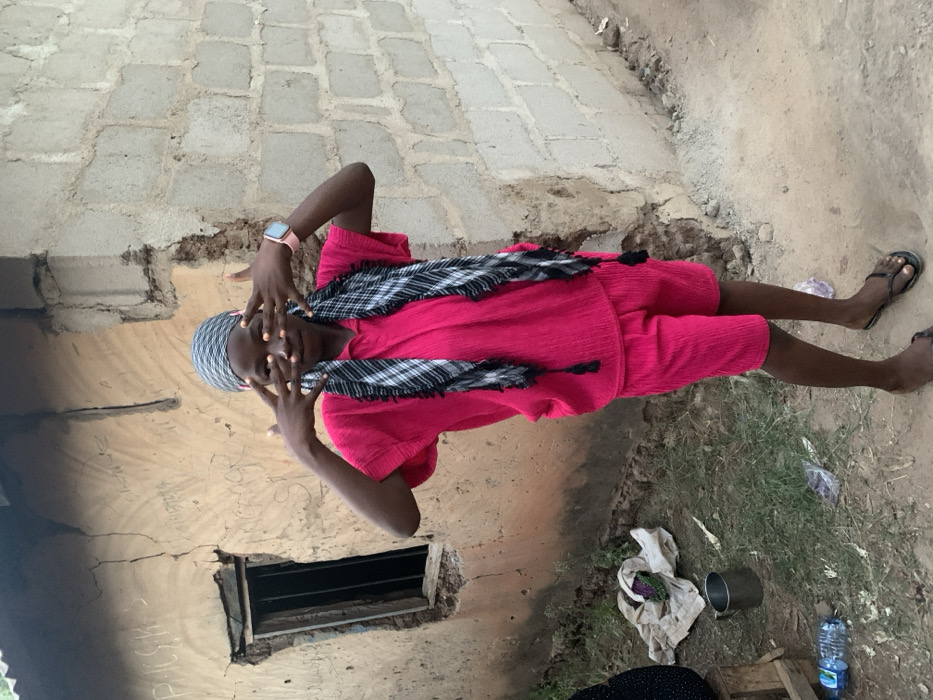 A person in a pink outfit poses with hands obscuring their face. They wear a patterned scarf and a watch. The background includes a weathered building with a small window, a brick or stone path, and an area with grass, a bottle of water, a tin cup, and cloth on the ground. The scene appears to be outdoors in a rural or less developed area.