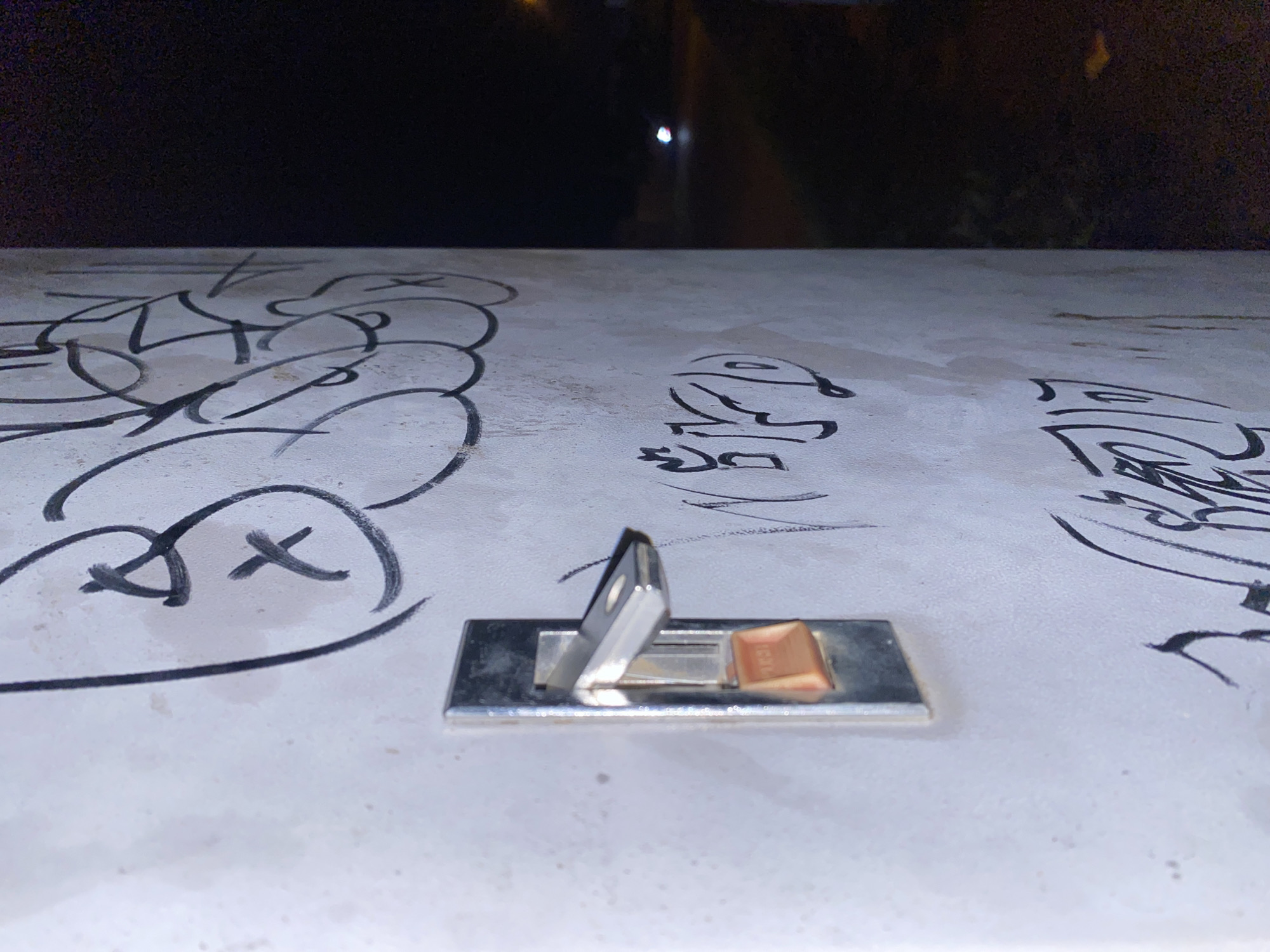 A close-up, side-profile shot captures a weathered, metallic light switch with an orange toggle against a rough, off-white concrete surface covered in black graffiti tags. The switch is positioned on the left side of the frame, appearing slightly tilted. The graffiti is dense, with large, stylized lettering dominating the concrete. To the right of the concrete surface, the scene transitions into darkness, revealing a hint of a dimly lit outdoor area with what appears to be a road or pathway and a distant, blurred light source, possibly a vehicle's headlights or a streetlamp. The overall impression is one of urban decay and nighttime ambiance. The context of Davao City, Philippines, suggests this scene could be found in various public or semi-public spaces, such as alleyways, under bridges, or along less maintained urban thoroughfares, where graffiti and worn infrastructure are common.