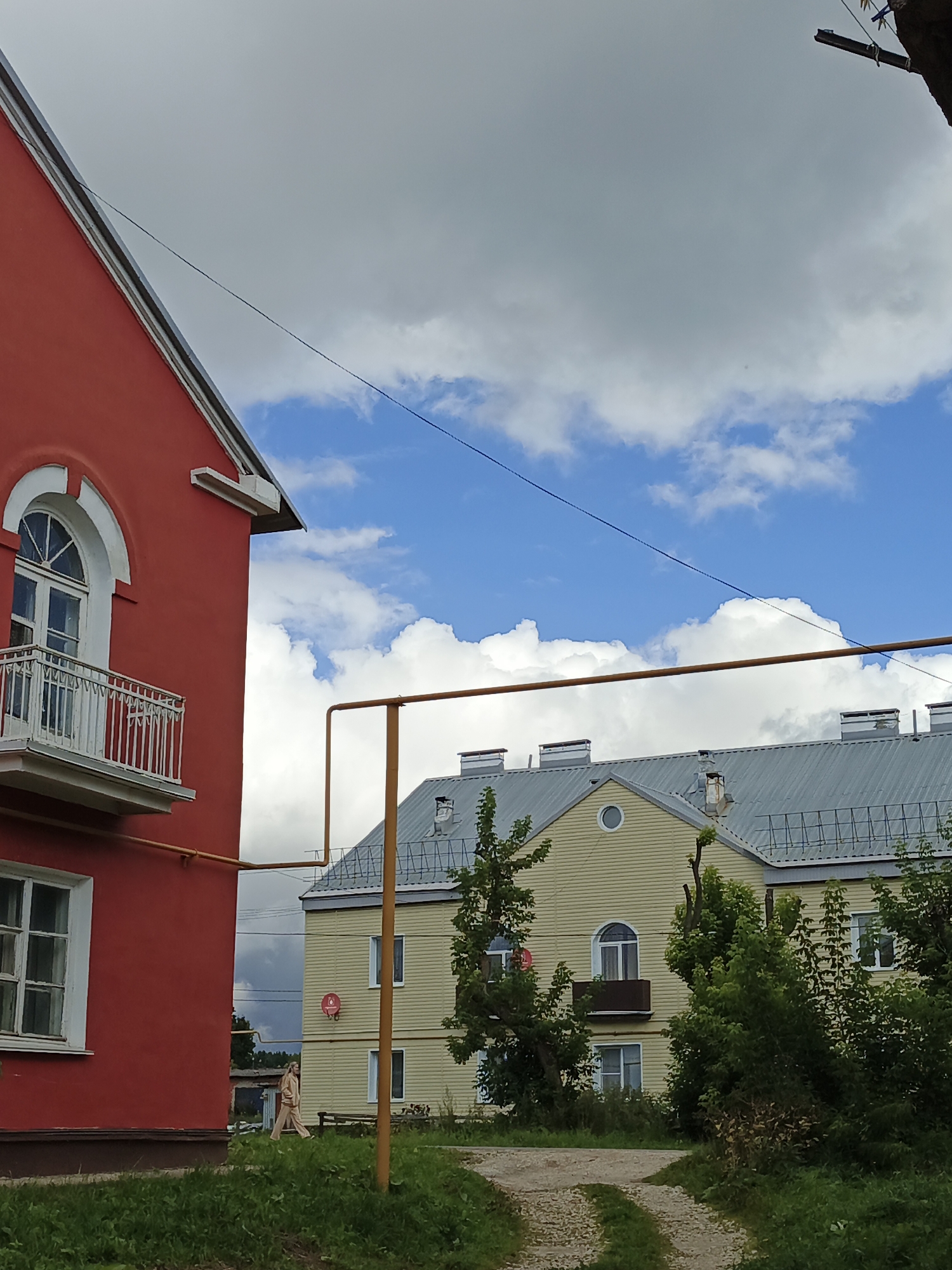 The image depicts an outdoor scene in Medvedyovka, Russia, featuring two residential buildings and a cloudy sky.
In the foreground, on the left, is a bright red building with a white-trimmed arched window and a small white balcony. A portion of a white-framed window is visible on the lower section of this building. A brown trim runs along the base of the red structure.
To the right of the red building is a light yellow, two-story residential building. It has a gray, corrugated metal roof with several dormer windows and what appear to be ventilation pipes or chimneys. The yellow building has multiple white-framed windows, and a small, dark-colored balcony protrudes from the upper level. A circular decorative element is present above the upper floor. A red satellite dish is mounted on the exterior wall.
A yellow gas pipe runs horizontally across the mid-section of the image, connecting to a vertical support pole. Trees and overgrown green grass are visible in the foreground and around the buildings. A dirt path curves through the grassy area in front of the yellow building.
A lone individual, wearing light-colored, possibly beige or tan, loose-fitting clothing, walks from left to right in front of the yellow building.
The sky is a mix of bright blue patches and large, gray, and white clouds, indicating dynamic weather conditions. Power lines are faintly visible crossing the sky.