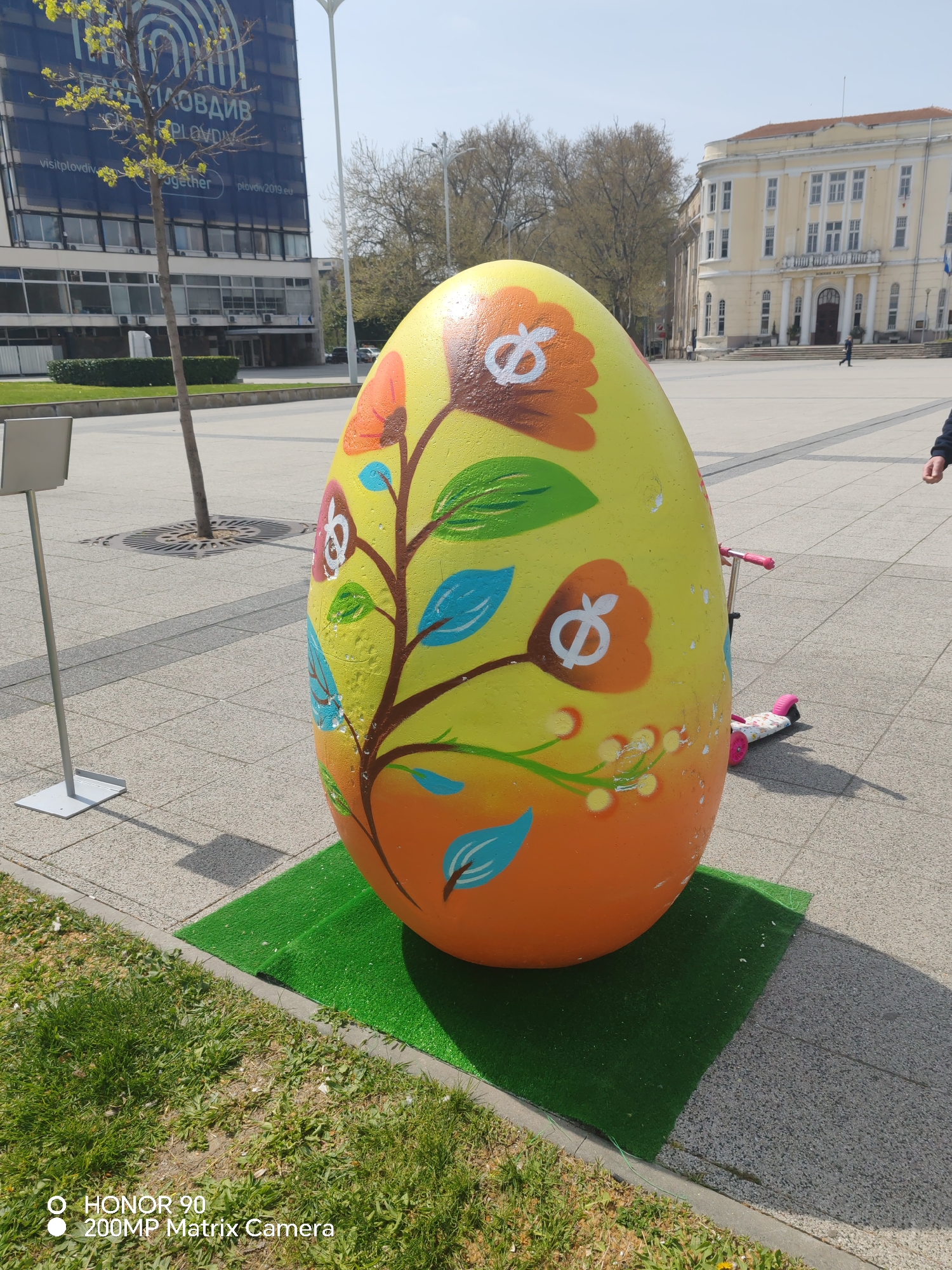 The image shows a large, egg-shaped sculpture decorated with floral designs in a public square. The egg is painted yellow and orange with flowers, stems, and leaves in various colors. It sits on a square of artificial green turf. In the background are buildings, including a modern office building with a banner that reads "ПЛОВДИВ PLOVDIV visitplovdiv together plovdiv2019.eu". A tan, classical-style building is also visible further back. A tree is in front of the office building, and other trees are visible near the classical building. The ground is paved with light gray paving stones. A metal sign stand is nearby on the left. The location appears to be in the city of Plovdiv, Bulgaria, based on the text on the banner. A scooter is visible behind the egg, and a person's arm is partially visible to the right.